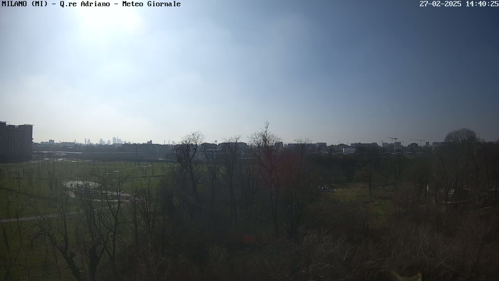 A clear, sunny day reveals a cityscape in the distance, partially obscured by a foreground of bare winter trees and a grassy field.