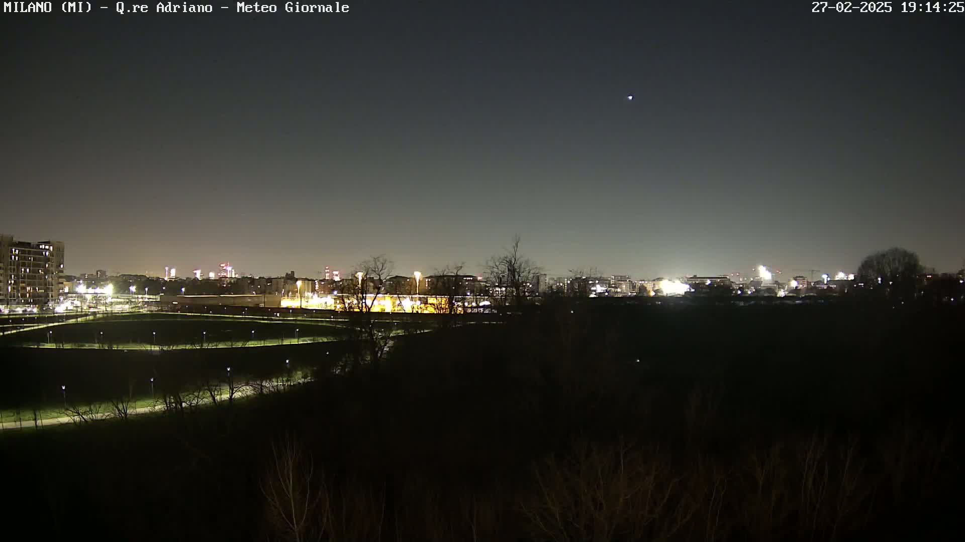 A nighttime view of a city skyline across a dark, sparsely vegetated area, under a clear sky.