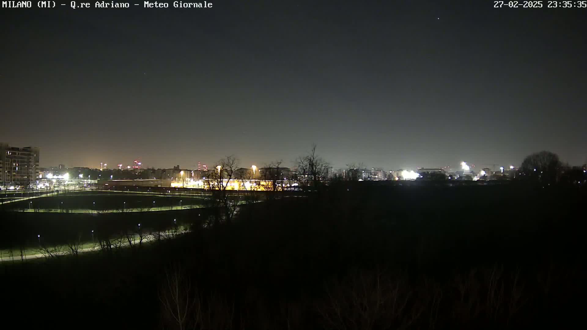 A nighttime view of a city skyline across a dark, sparsely lit park under a clear, dark sky.