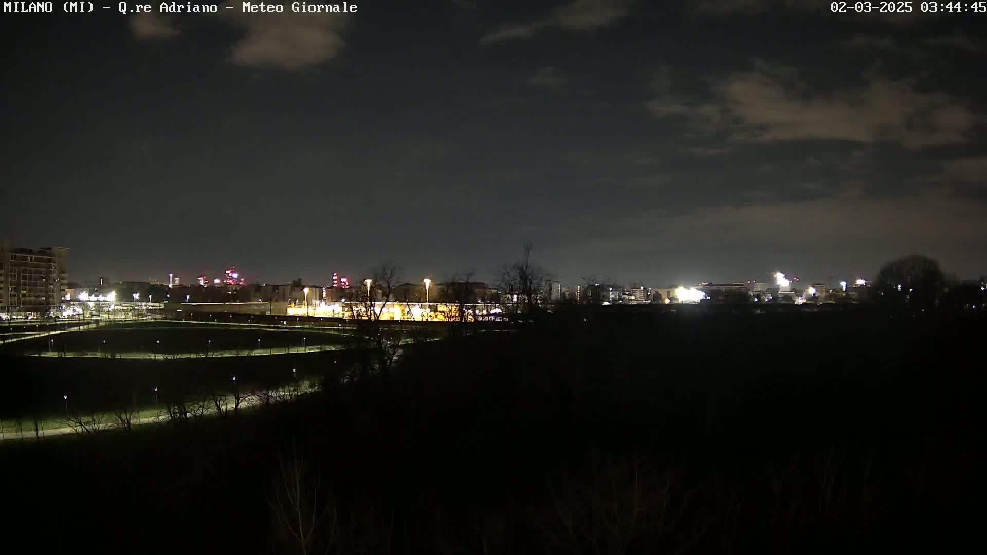A nighttime view of a city skyline under a partly cloudy sky, with illuminated paths and dark trees in the foreground.
