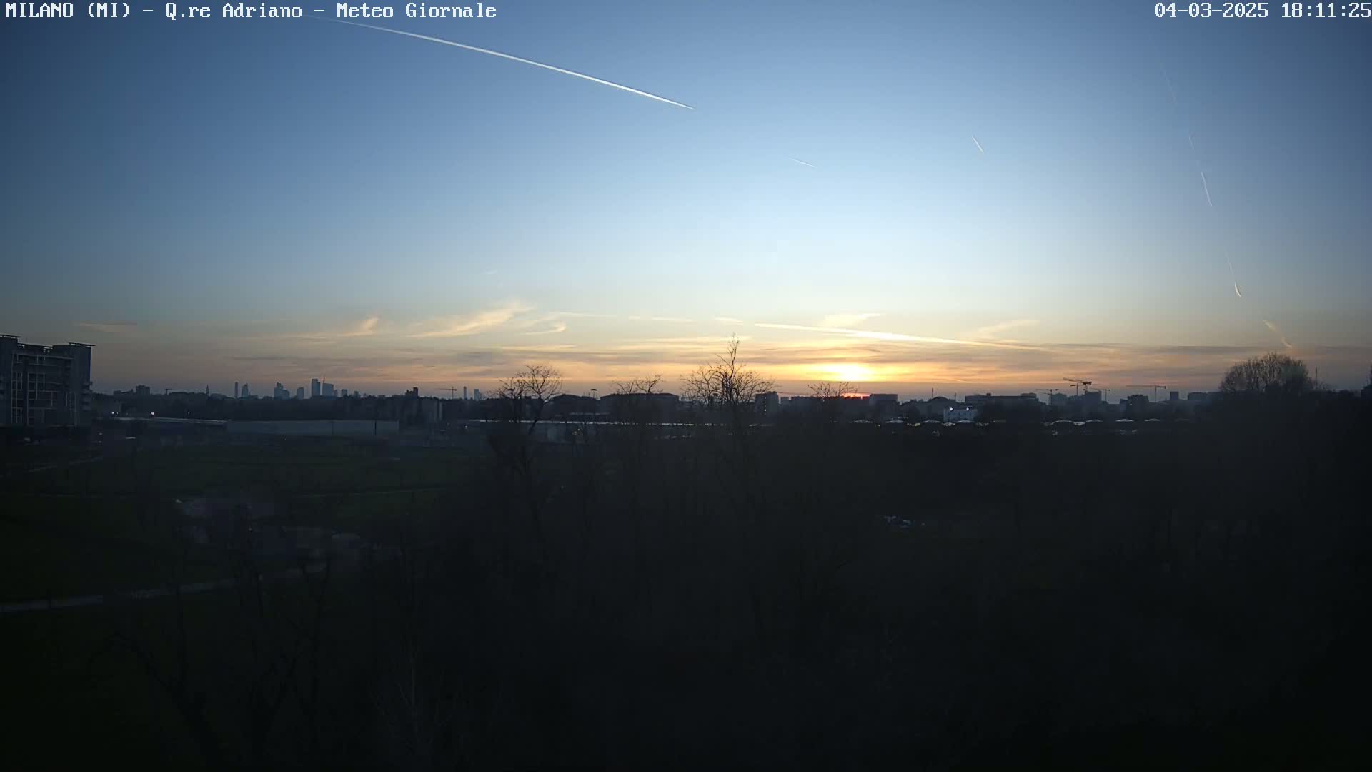 A city skyline at sunset, under a clear sky with contrails, is viewed from across a field of dark trees and grass.