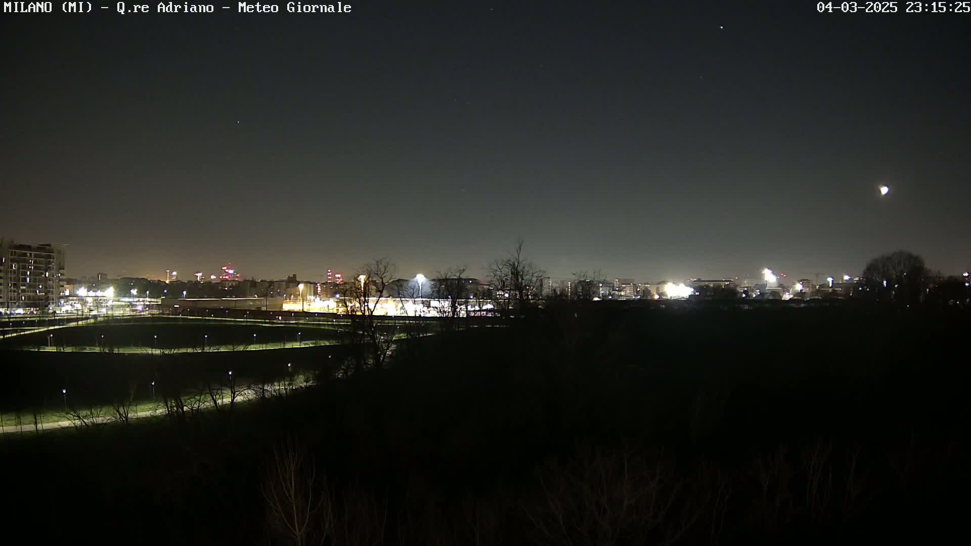 A nighttime view of a city skyline and a dark, tree-covered area in the foreground, under a clear, starry sky with a bright moon visible.