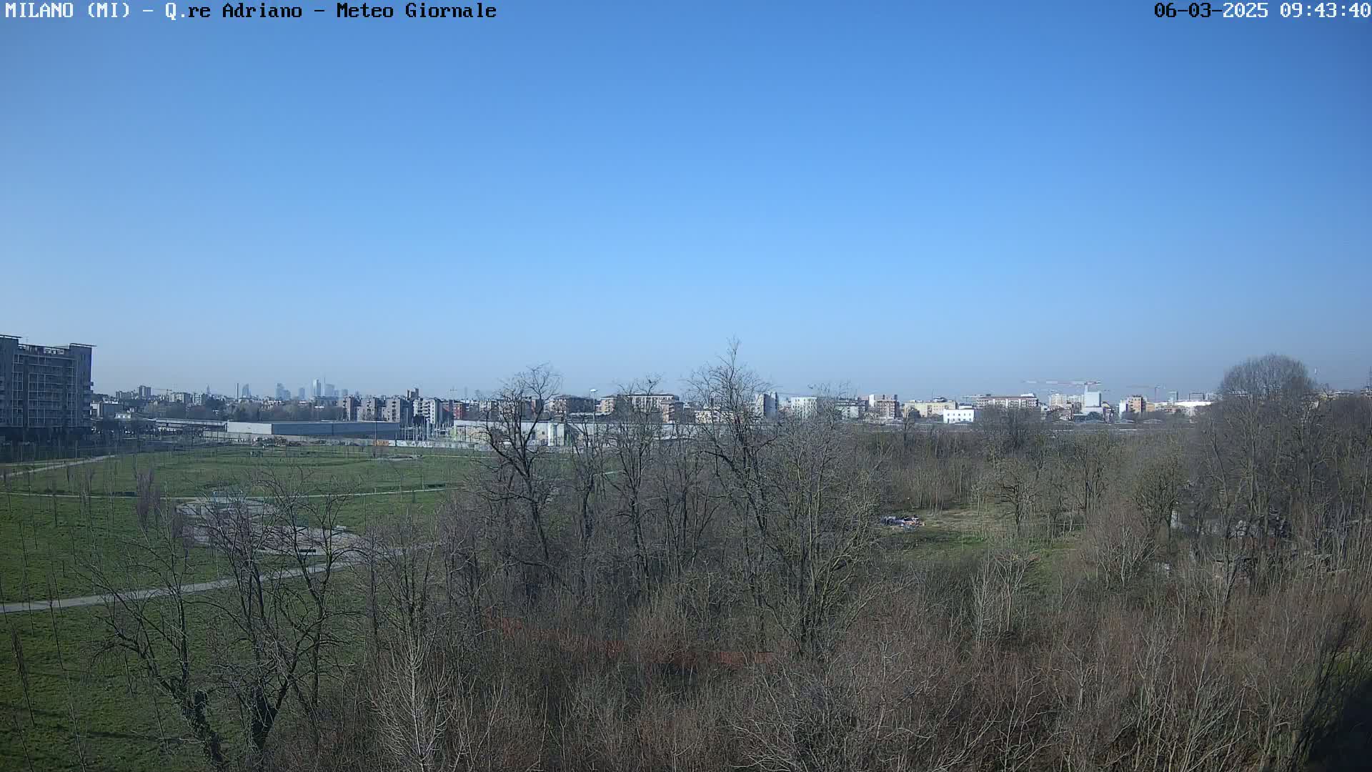 A clear blue sky overlooks a park with bare trees and grassy areas, beyond which a city skyline is visible in the distance.