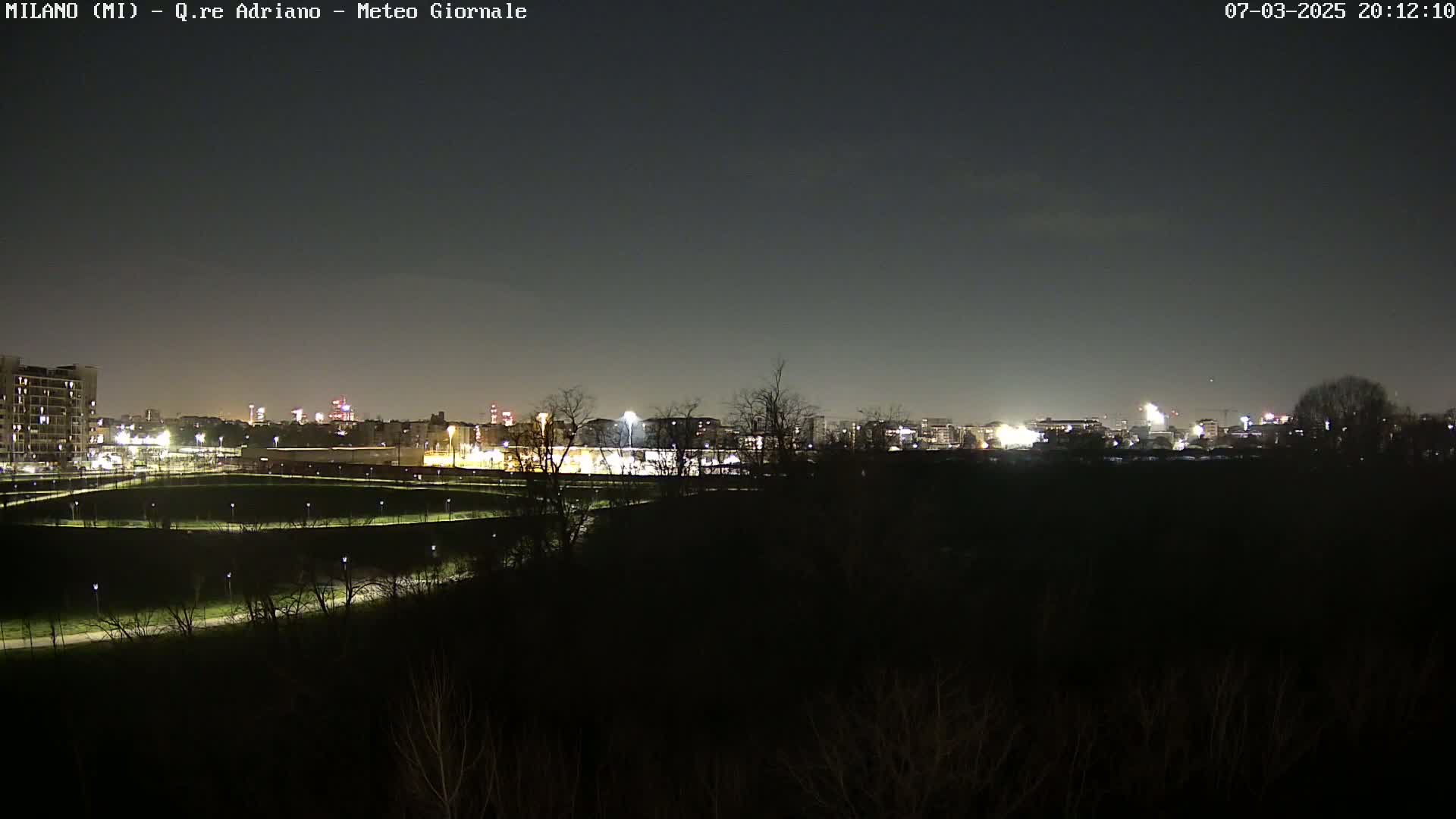 A nighttime view of a city skyline illuminated with lights, seen from across a dark, empty field under a clear, dark sky.