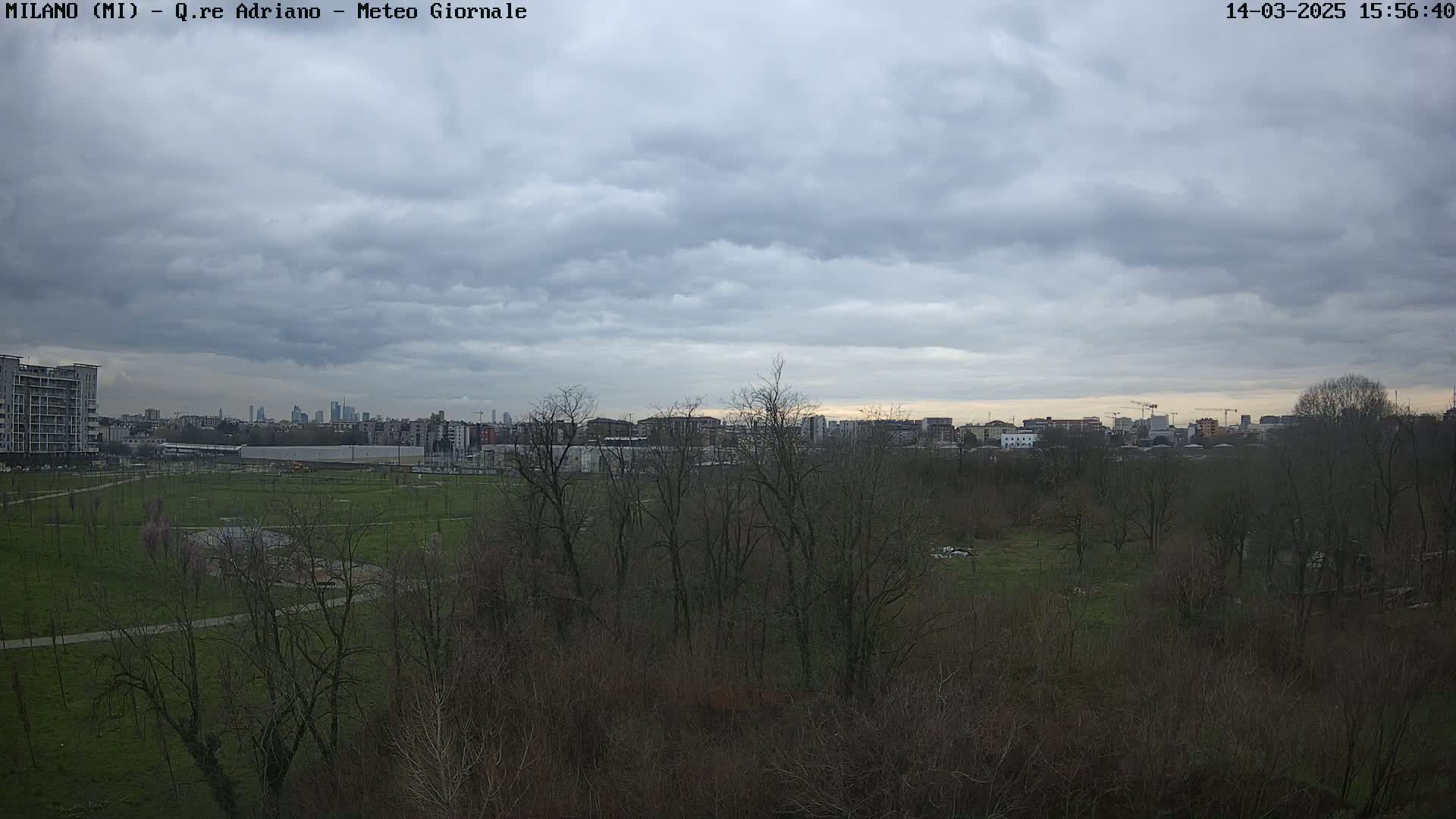 A city skyline is visible in the distance behind a sprawling park with bare trees under a cloudy sky.