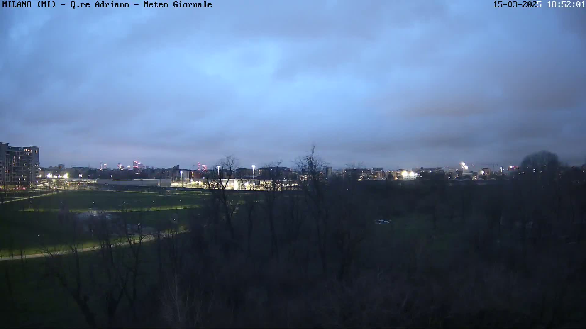A city skyline is visible at dusk under a mostly cloudy sky, beyond a dark, tree-lined park with a lit field.