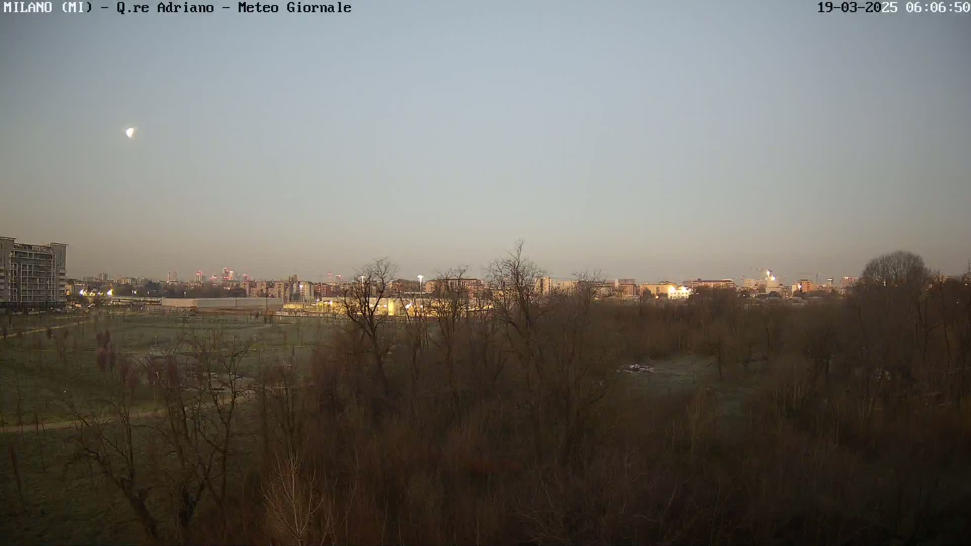 A pale, pre-dawn sky shows a crescent moon above a flat landscape of bare trees and a distant city skyline.