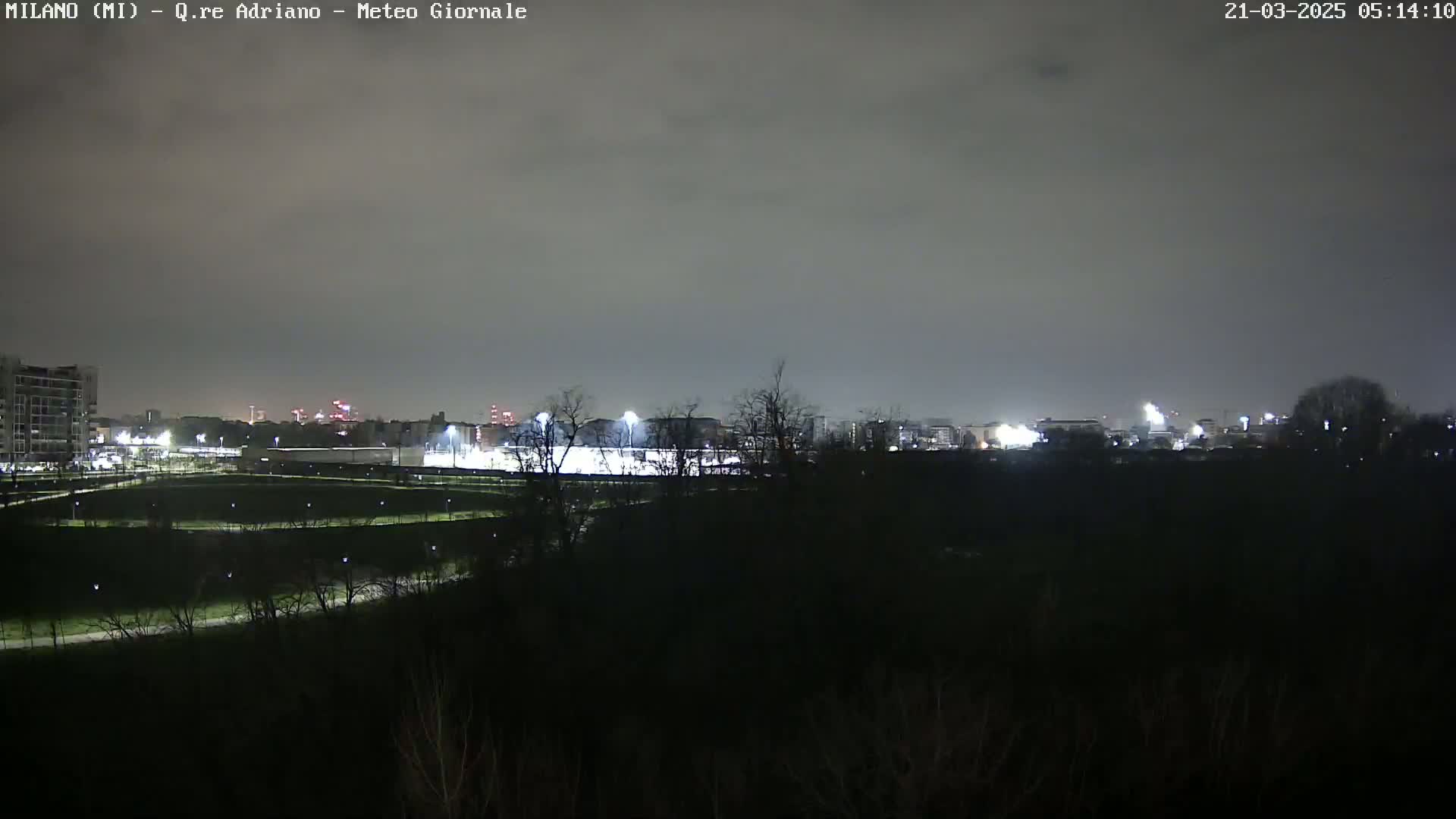 A nighttime view of a park with illuminated buildings and fields in the distance under a cloudy sky.
