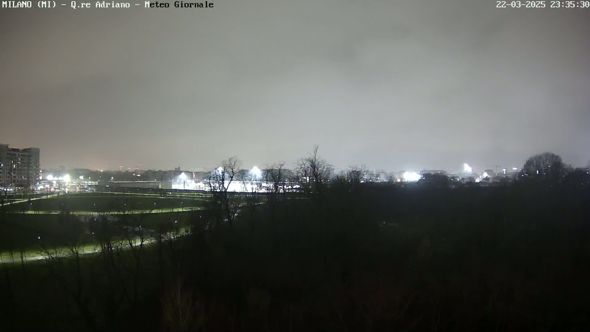 A nighttime view of a city park with lights illuminating a playing field and buildings in the distance under a cloudy sky.