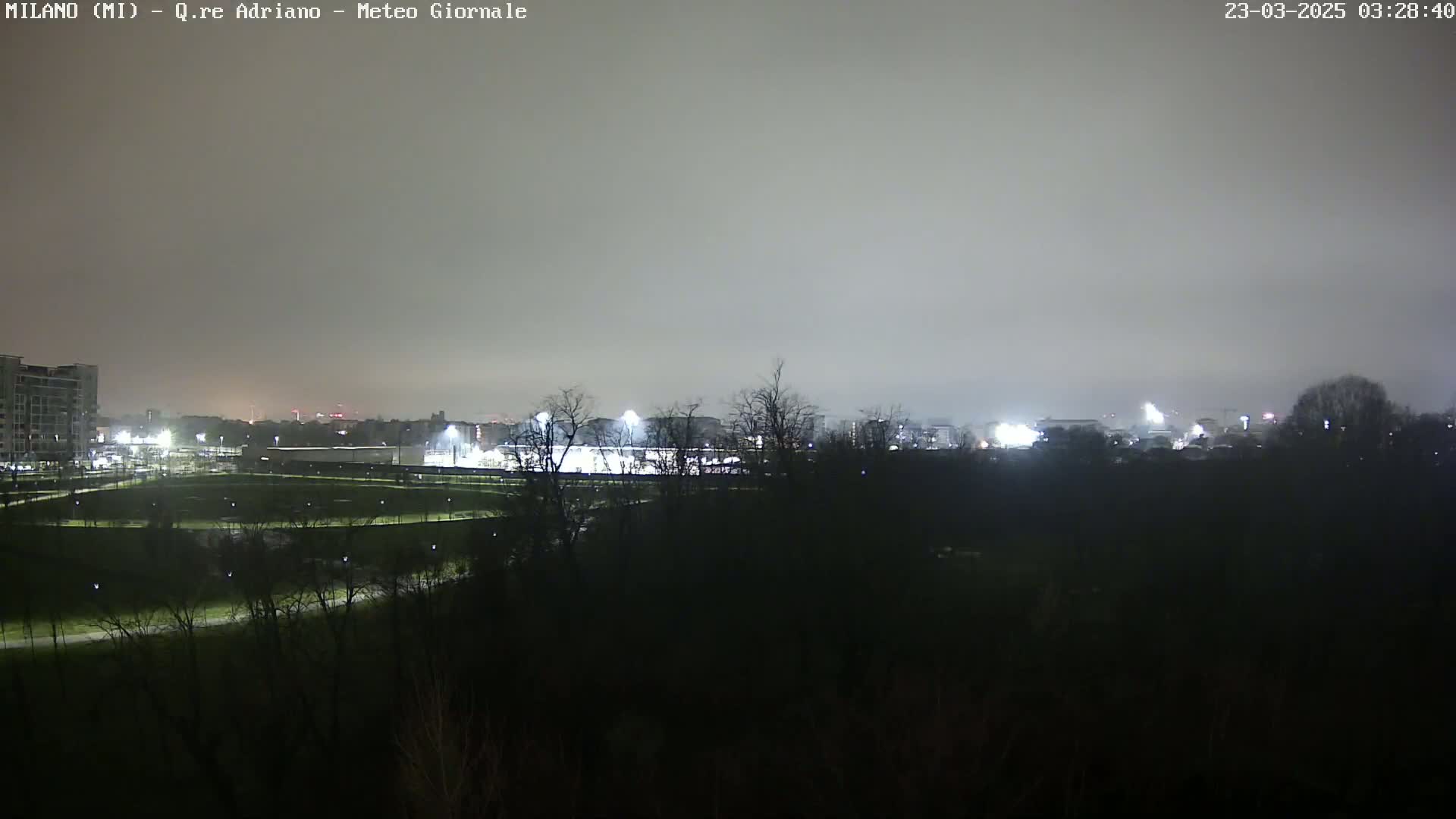 A nighttime view of a park and sports field, illuminated by artificial lights under a hazy sky.