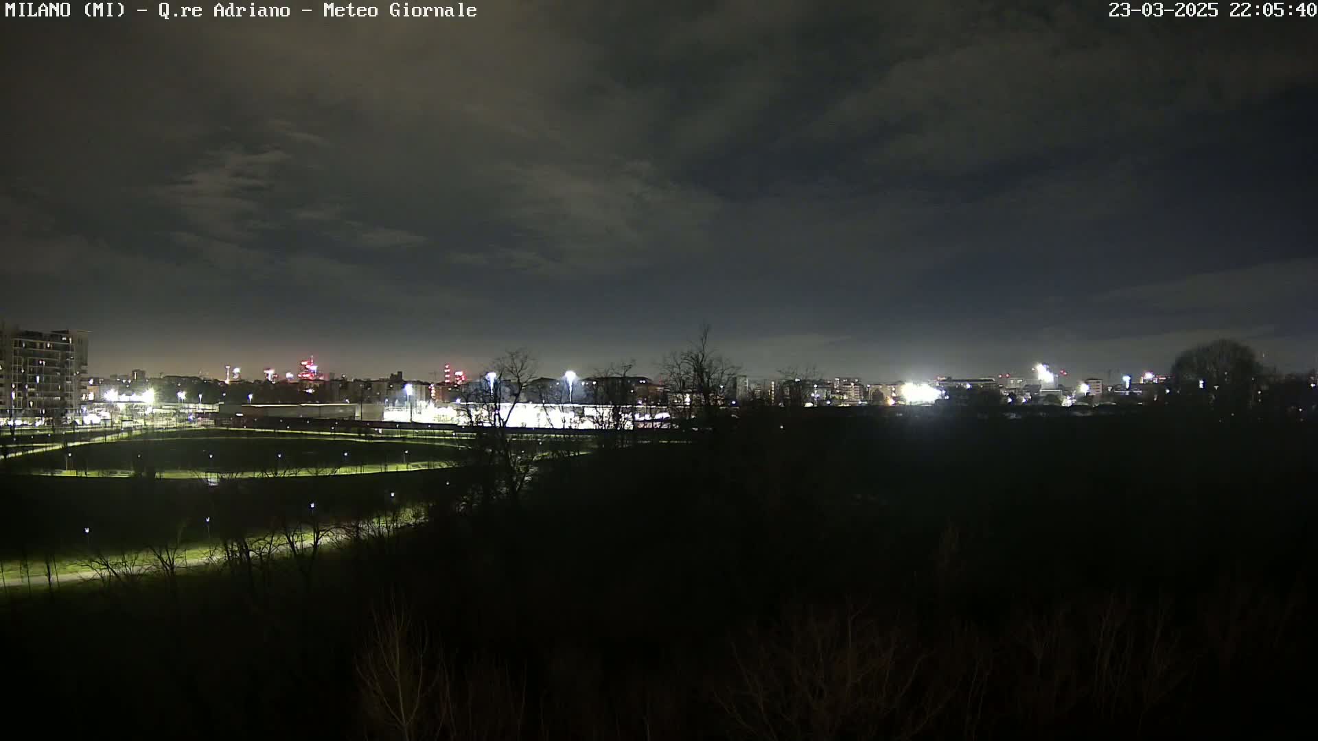 A nighttime view of a city skyline across a dark, grassy field under a cloudy sky.