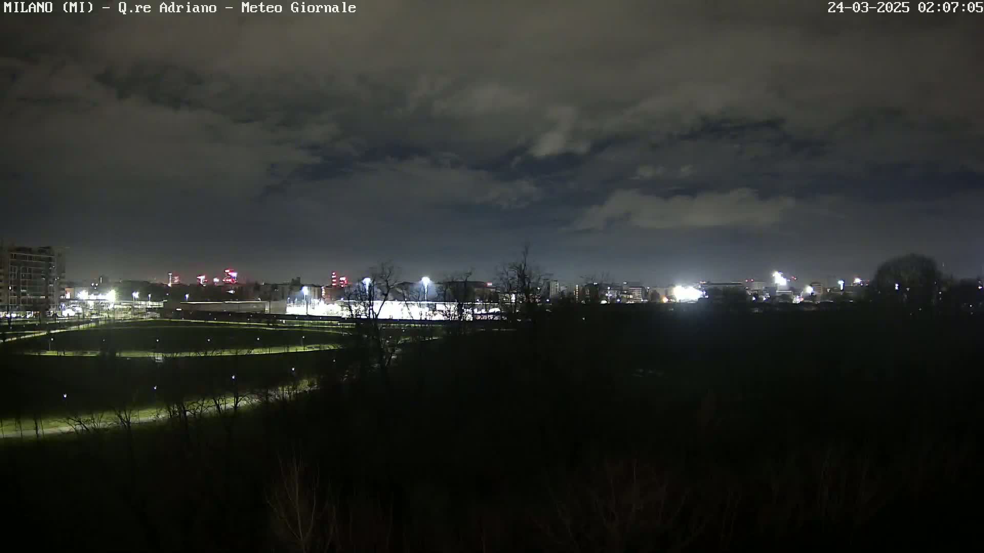 A nighttime view of a city skyline with a brightly lit field in the foreground under a cloudy sky.
