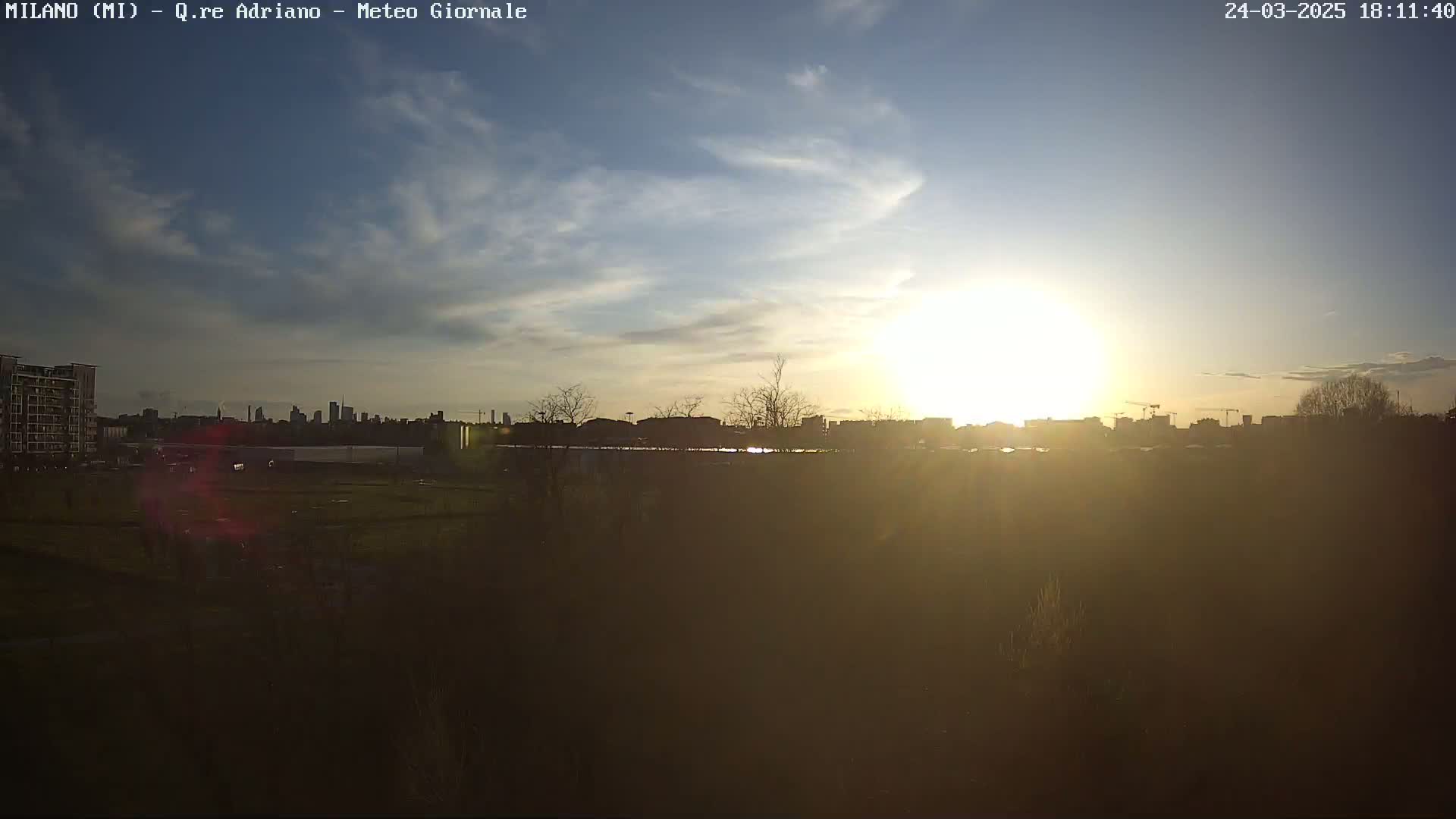 A partly cloudy sunset over a city skyline, viewed from a distance across a flat, grassy area.