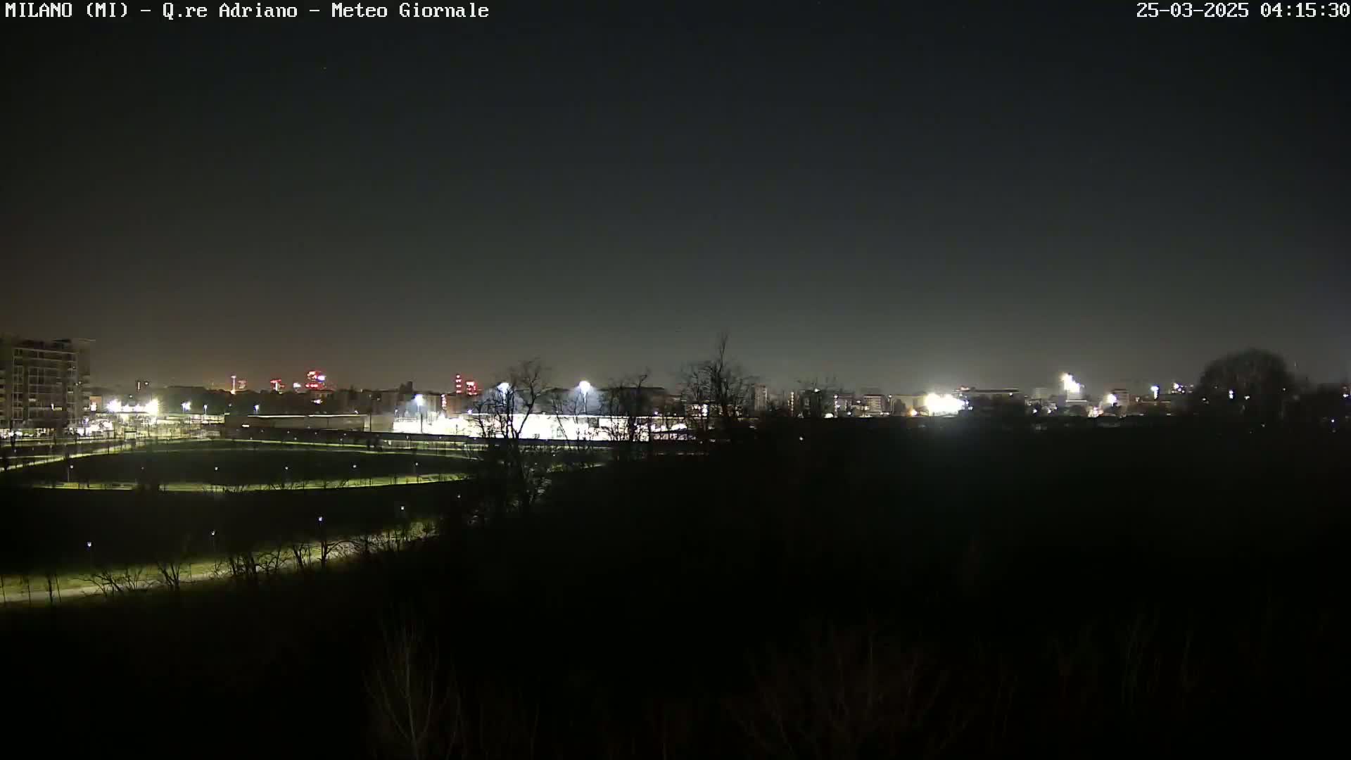 A nighttime long shot shows a dimly lit park with pathways, and a brightly lit sports field in the background, all under a dark, clear sky.