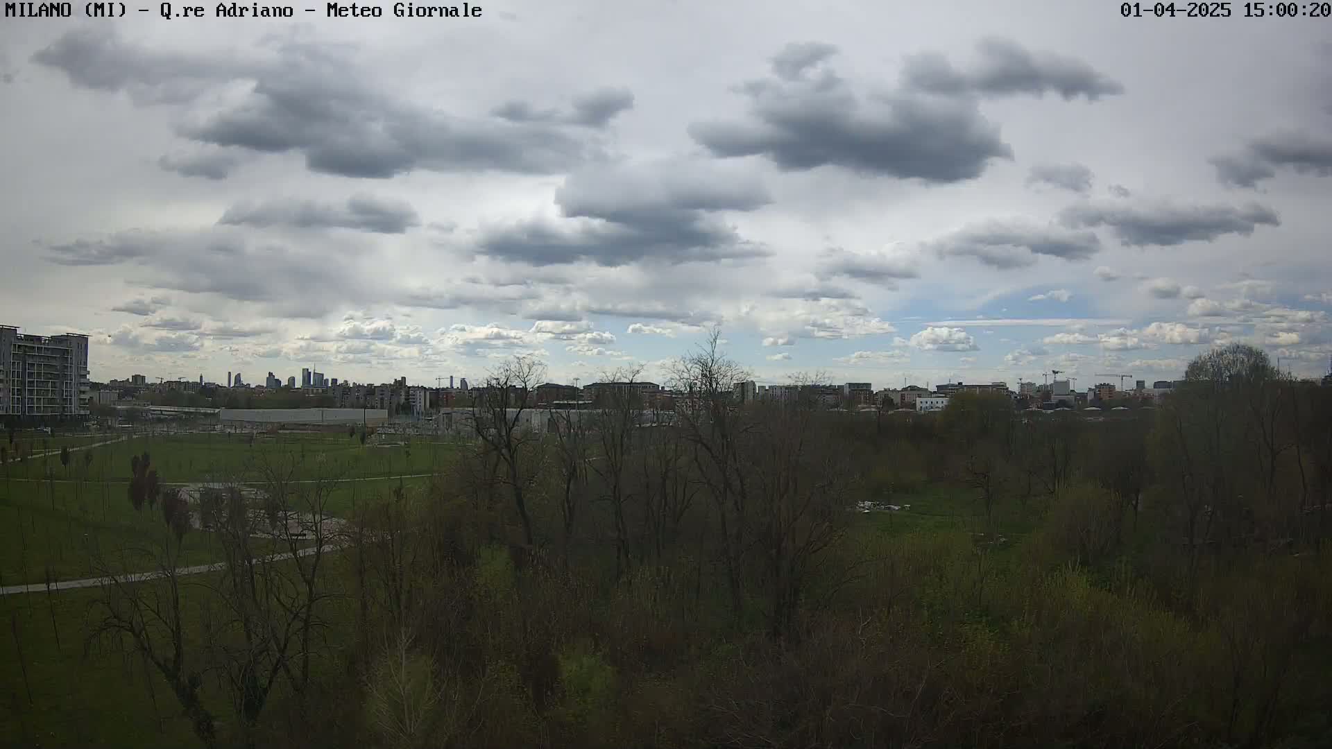 A partly cloudy sky overlooks a cityscape in the distance, beyond a sprawling park and wooded area.