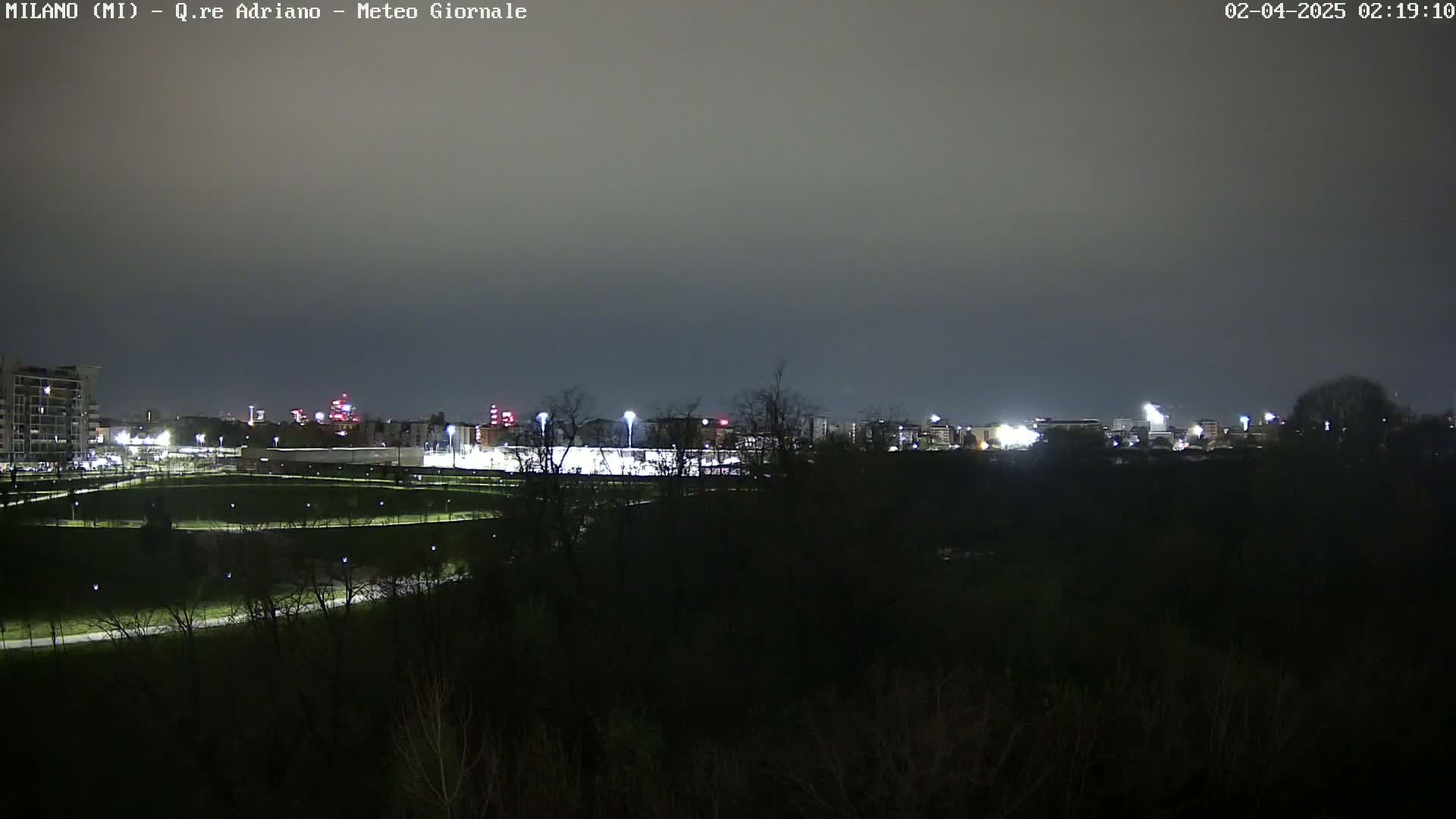A nighttime view shows a city skyline illuminated by lights across a dark, overcast sky, beyond a mostly dark expanse of trees and a lit-up field.