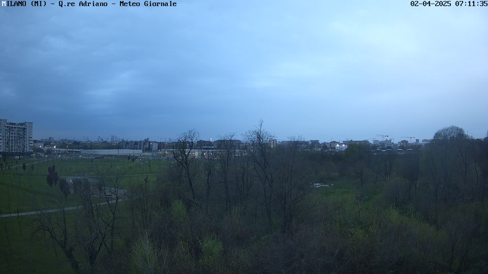 A city skyline is visible in the distance beyond a park with bare trees under a twilight sky.