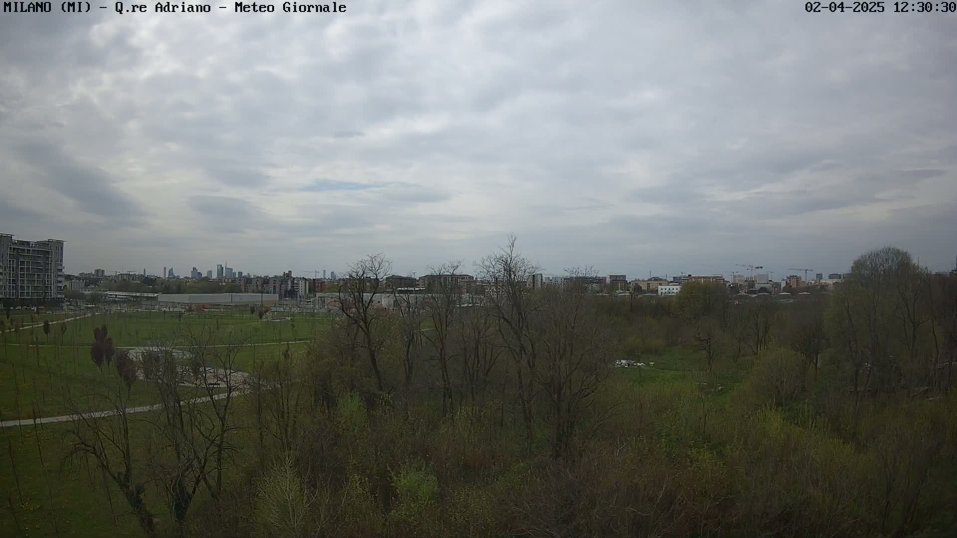 A mostly cloudy sky overlooks a park with budding trees and buildings in the distance.