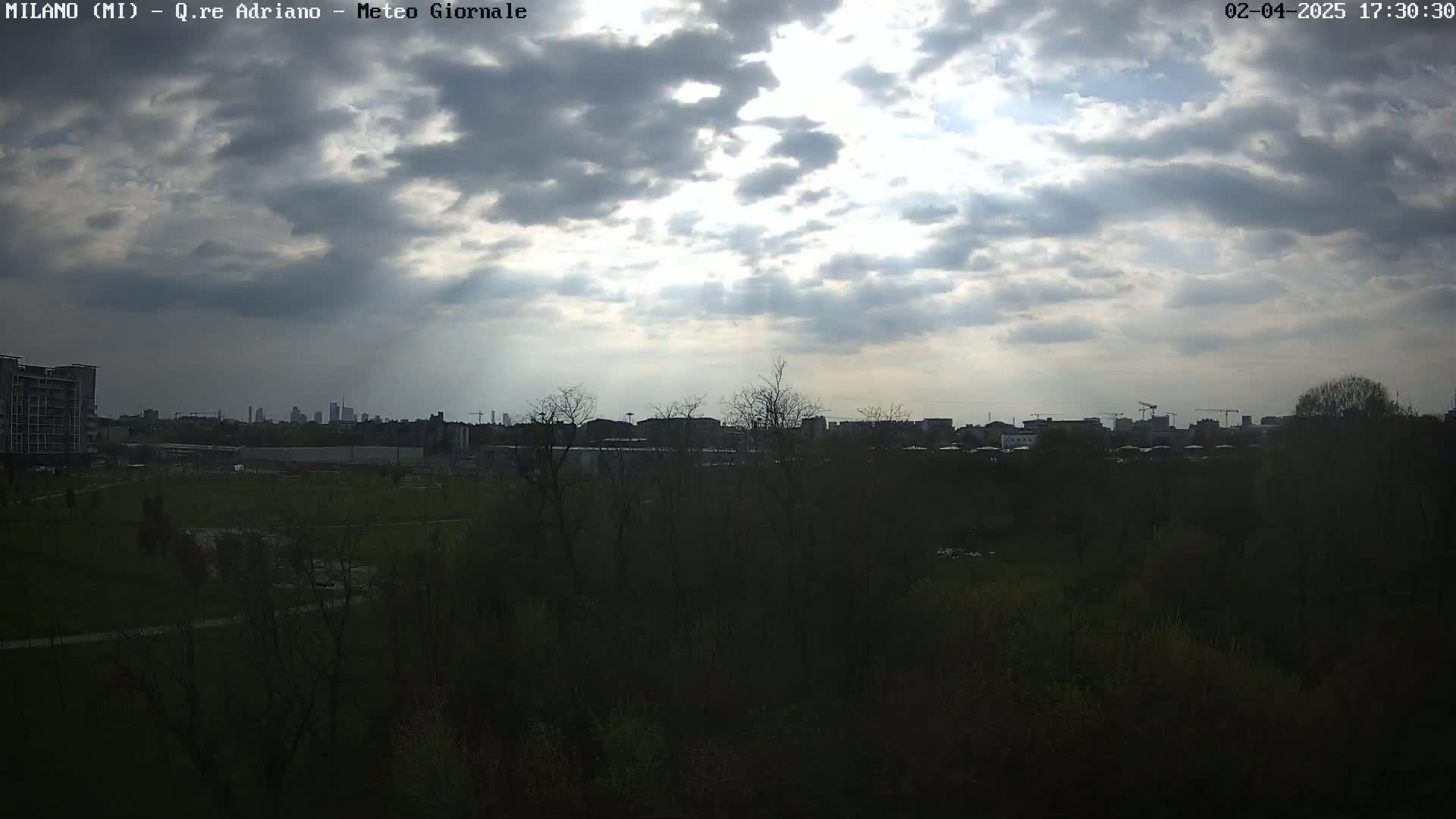 A partly cloudy sky shines over a city skyline visible beyond a dark, tree-lined park.
