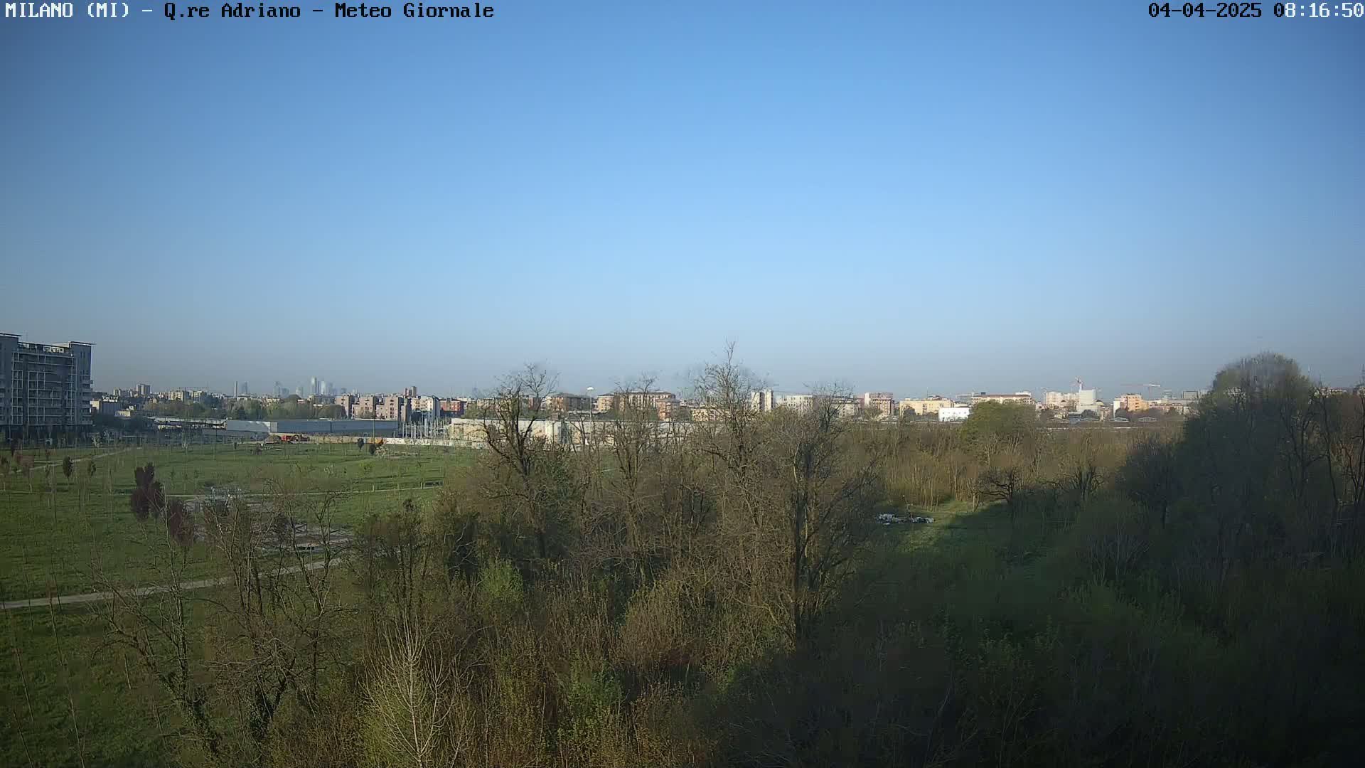 A clear, sunny day reveals a cityscape in the distance, partially obscured by a foreground of leafless trees and a grassy field.