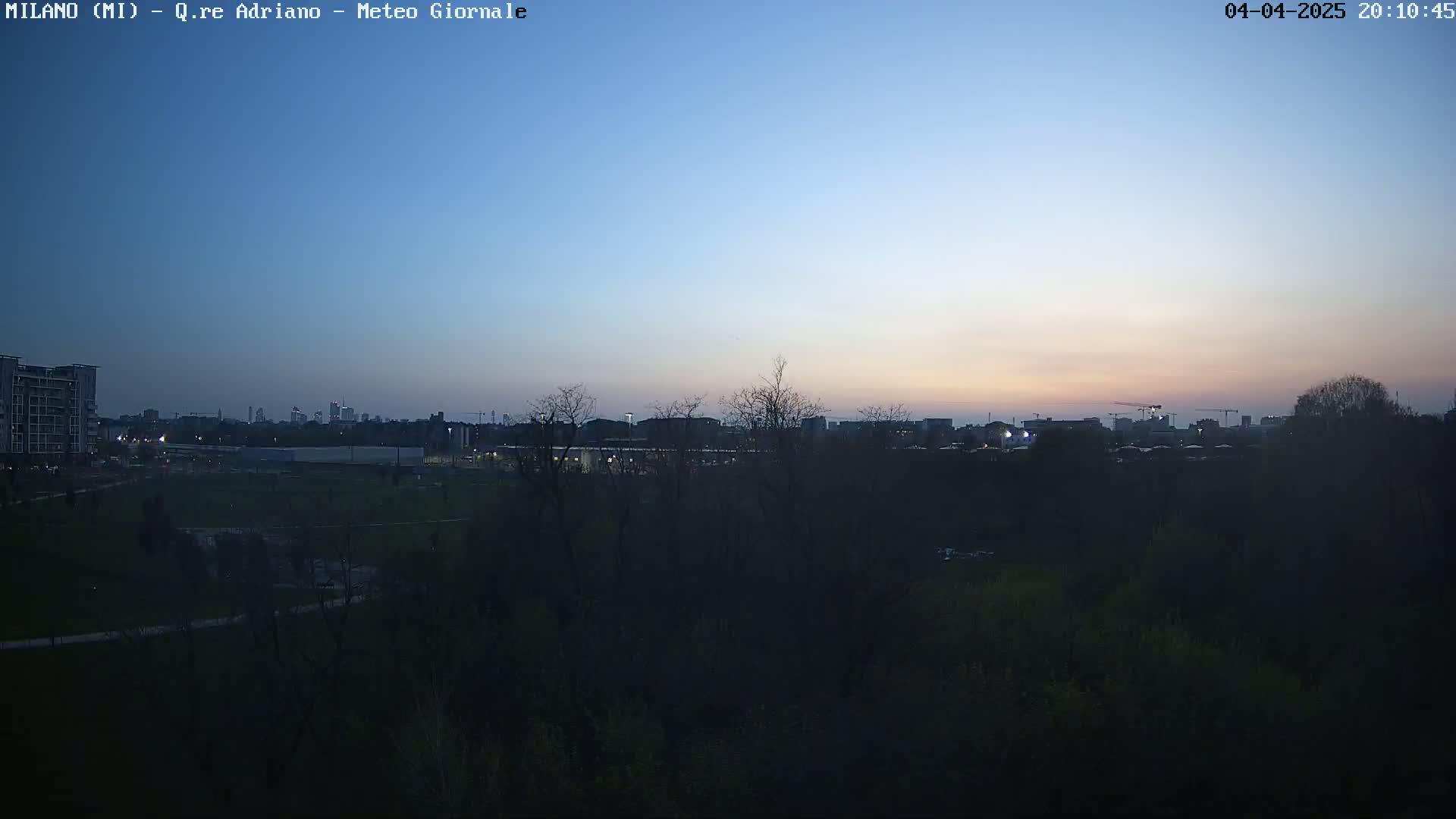 A twilight view shows a city skyline in the distance beyond a dark, tree-lined park.
