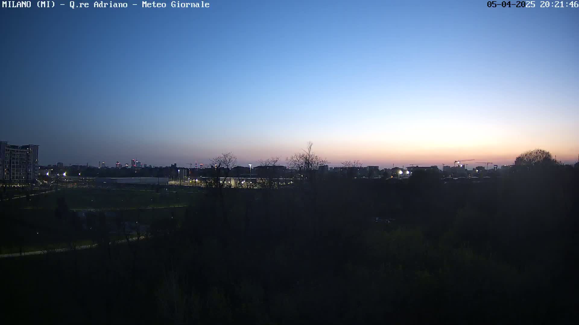 A twilight cityscape is visible beyond a dark, tree-lined foreground, with a clear, mostly blue sky.