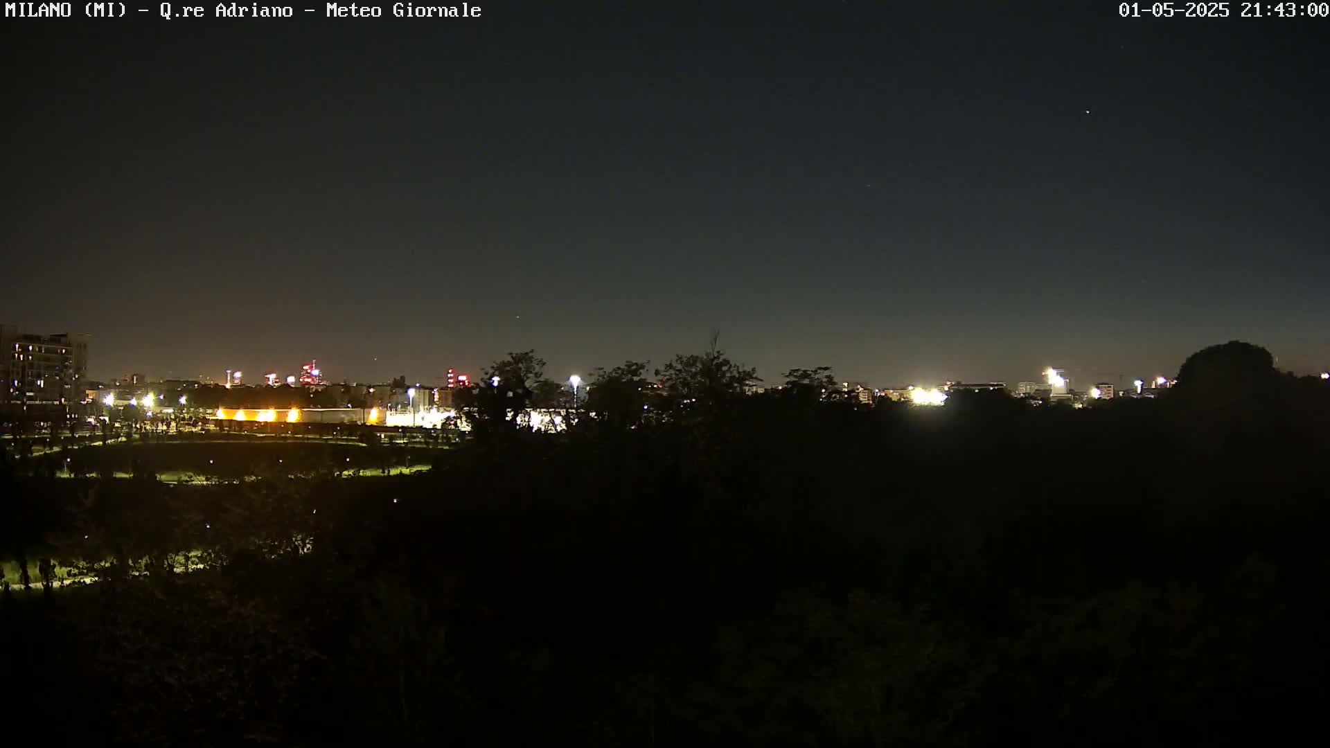 A nighttime view shows a city skyline dimly lit beyond a dark, tree-lined foreground under a clear, dark sky.