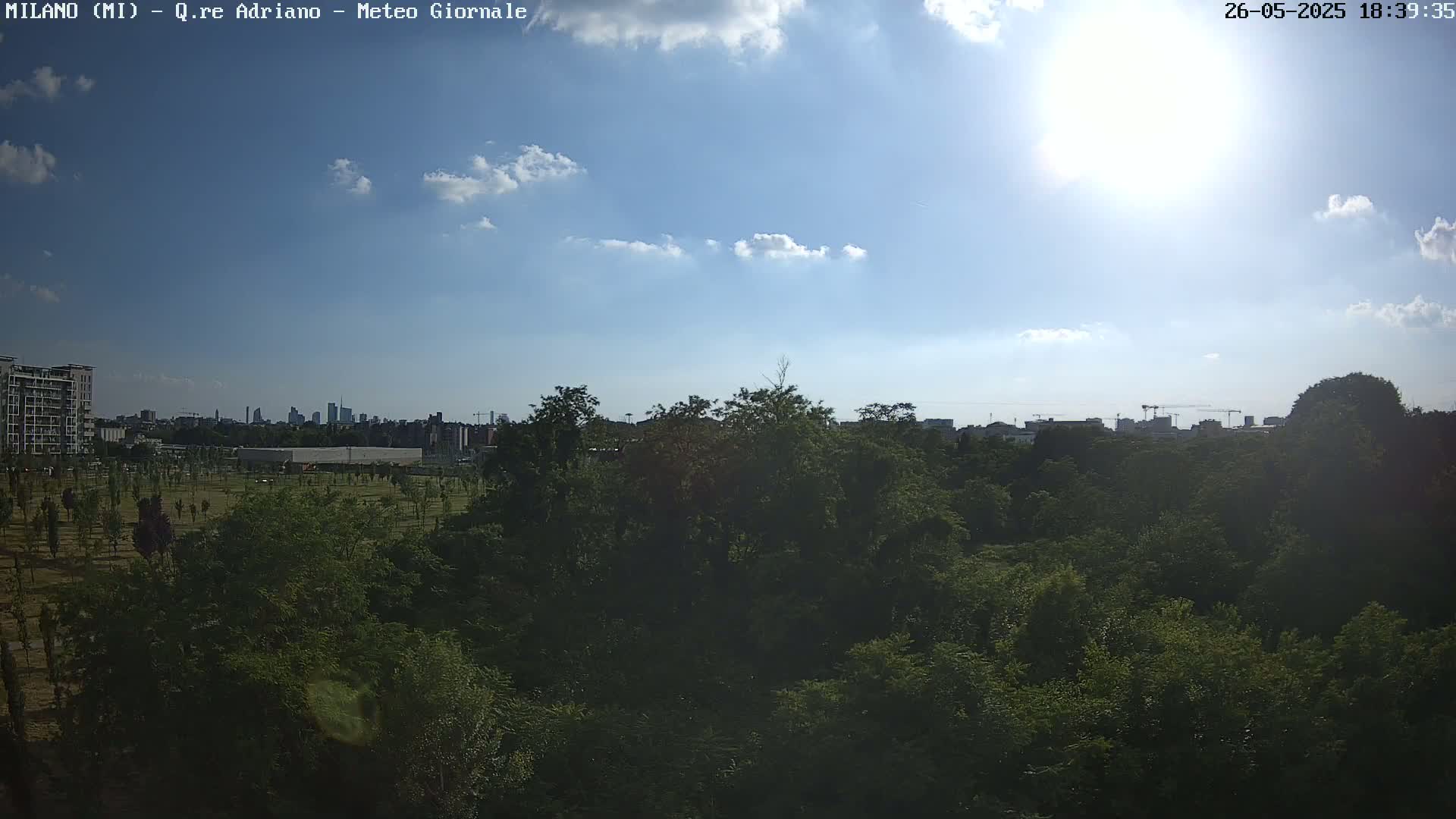 A sunny day with some clouds shows a view of a green, tree-filled park in the foreground, with a city skyline visible in the distance.