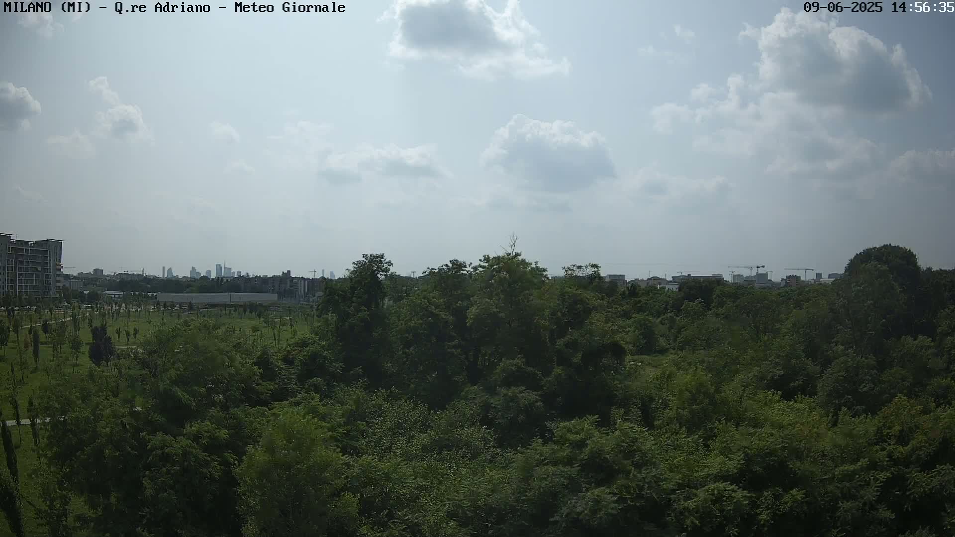 A partly cloudy sky overlooks a dense green park, with a distant cityscape visible on the horizon.