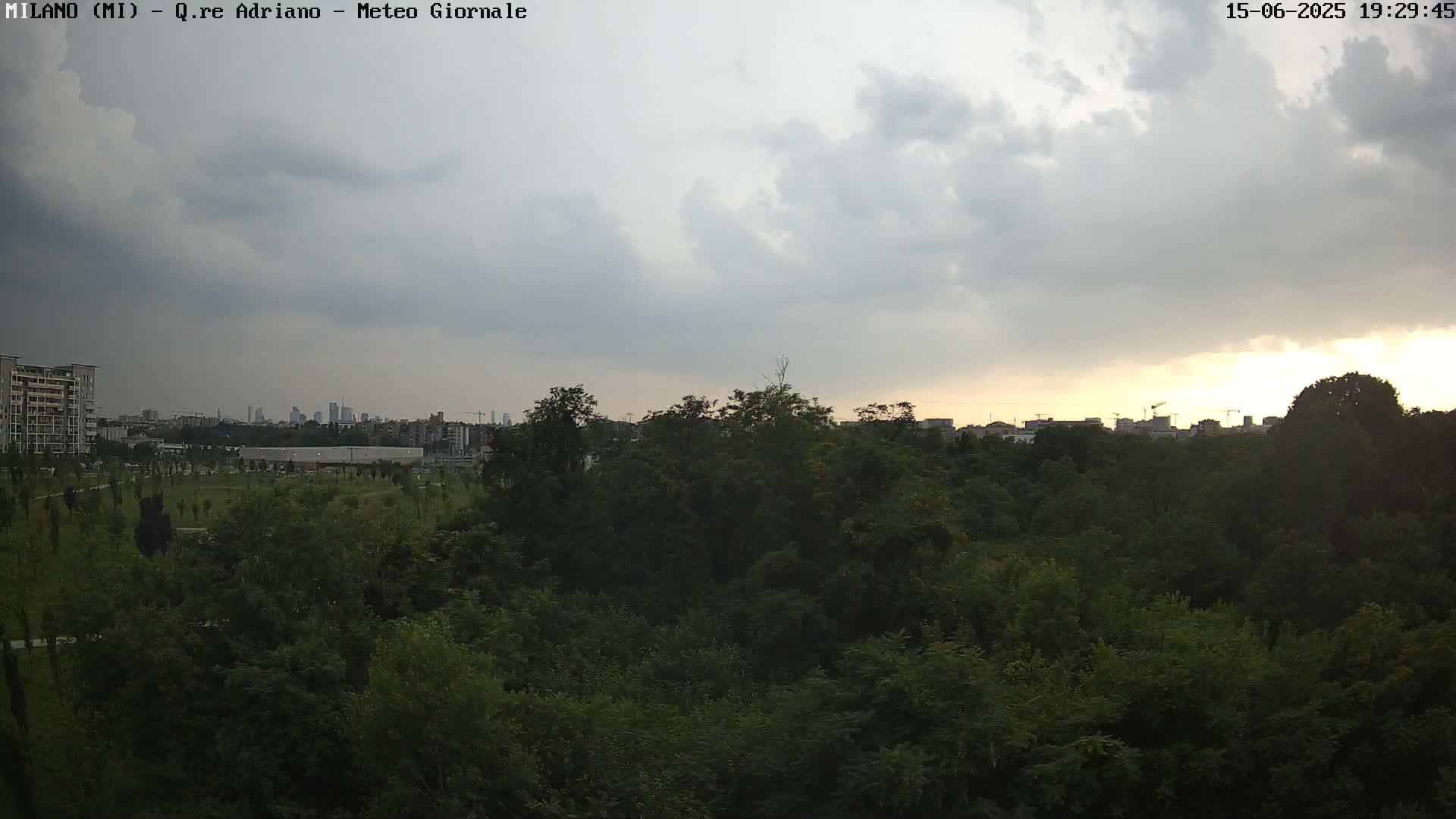 A mostly cloudy sunset over a dense green park, with a cityscape visible in the distance.