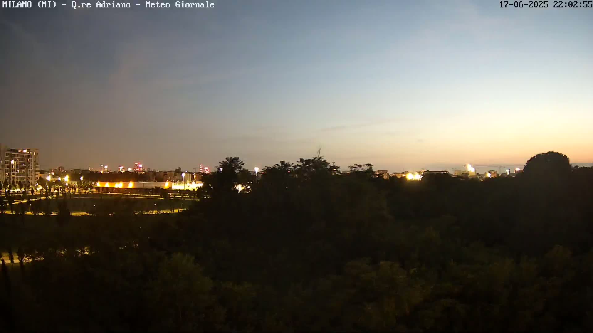A nighttime view of a city skyline with lights visible across a dark green expanse of trees under a twilight sky.