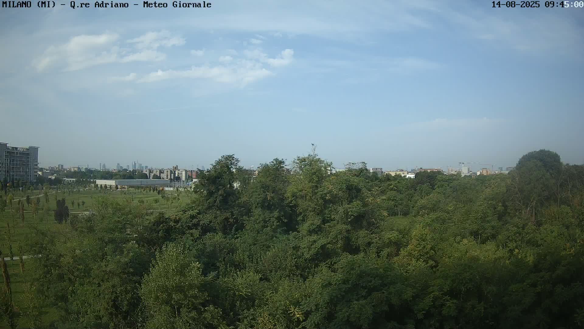A mostly sunny sky overlooks a dense green park with a city skyline visible in the distance.