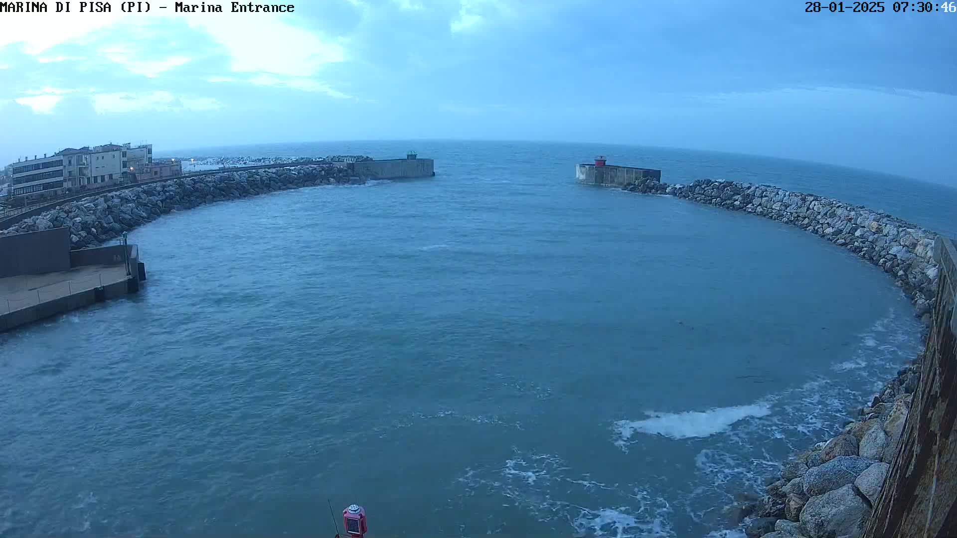 A harbor entrance with choppy water under an overcast sky is partially enclosed by rock walls and a small building.