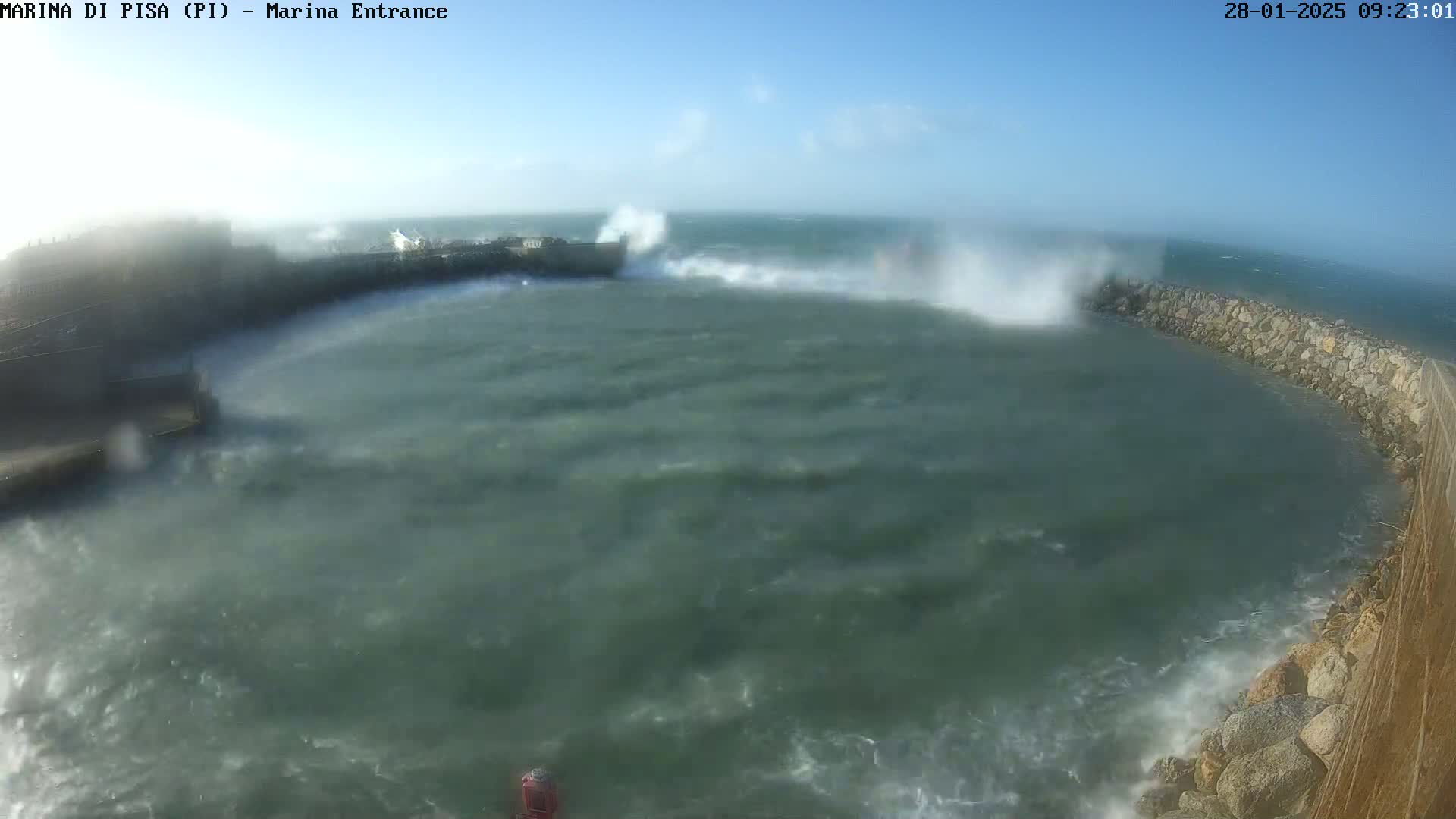 Large waves crash against a harbor wall under a bright, sunny sky.