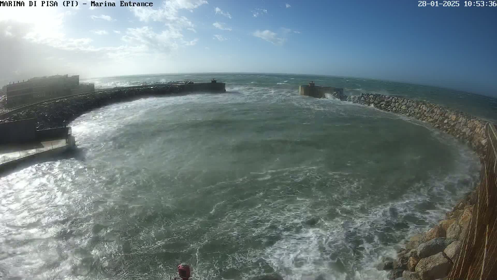 Rough, choppy waves crash against a stone harbor wall under a partly cloudy sky.