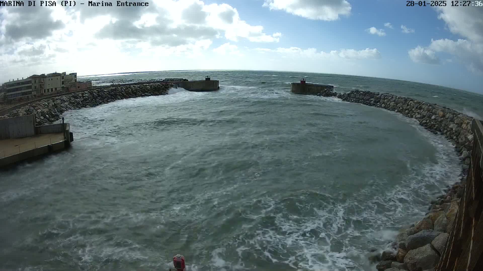 Rough, choppy water crashes against a rocky breakwater surrounding a harbor entrance under a partly cloudy sky.