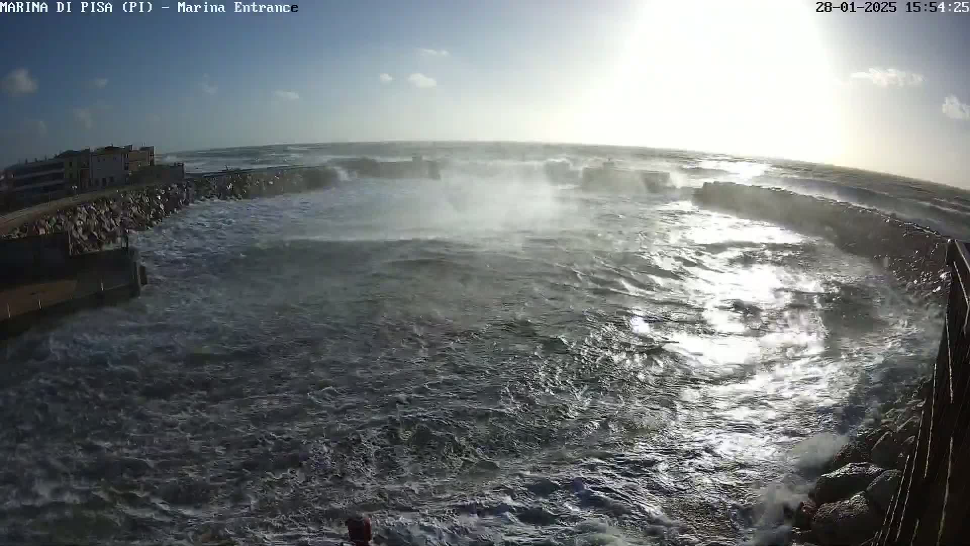 Rough, stormy seas crash against a harbor wall under a partly cloudy sky.
