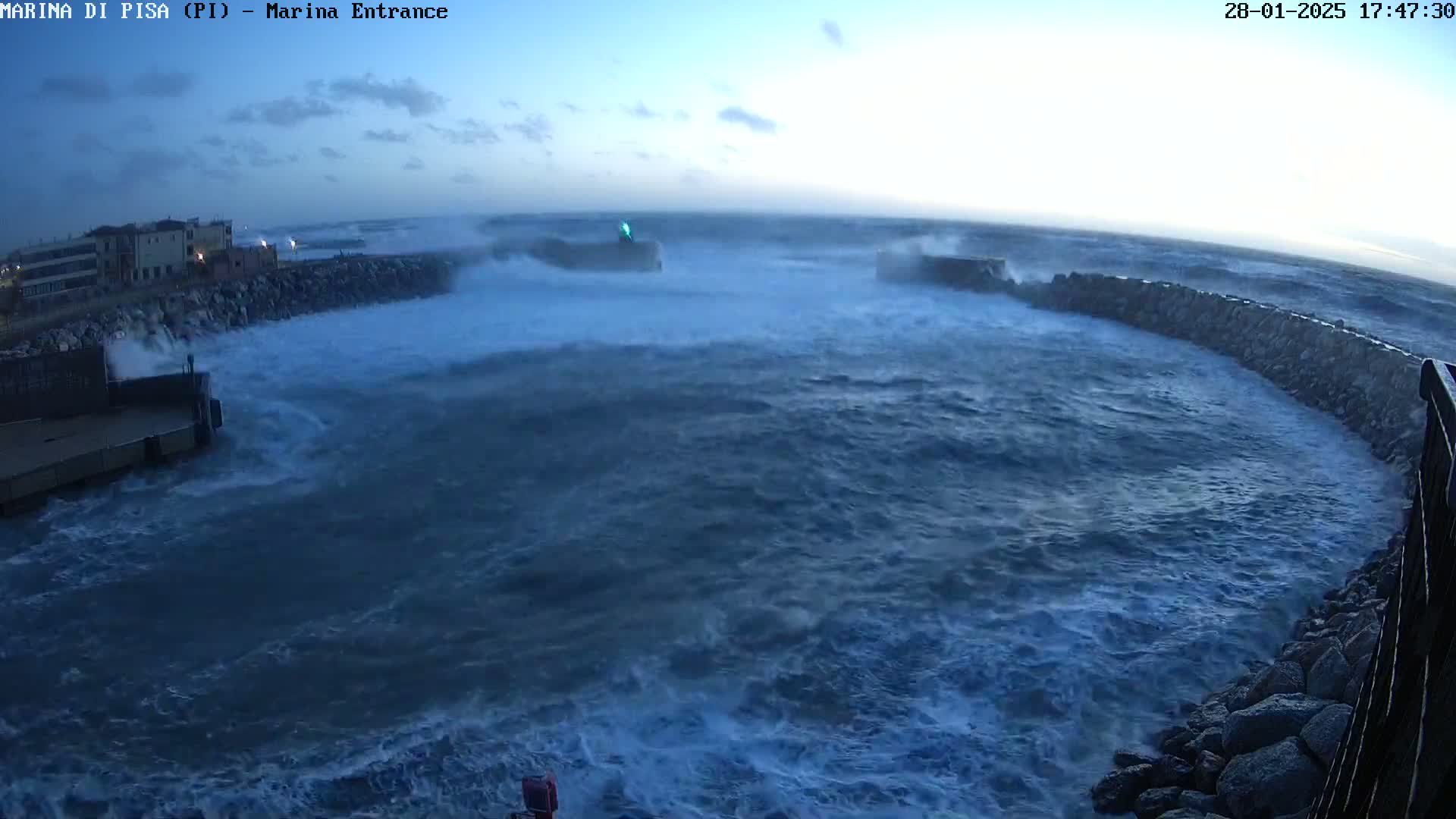 Large waves crash against a stone seawall near a building under a twilight sky.