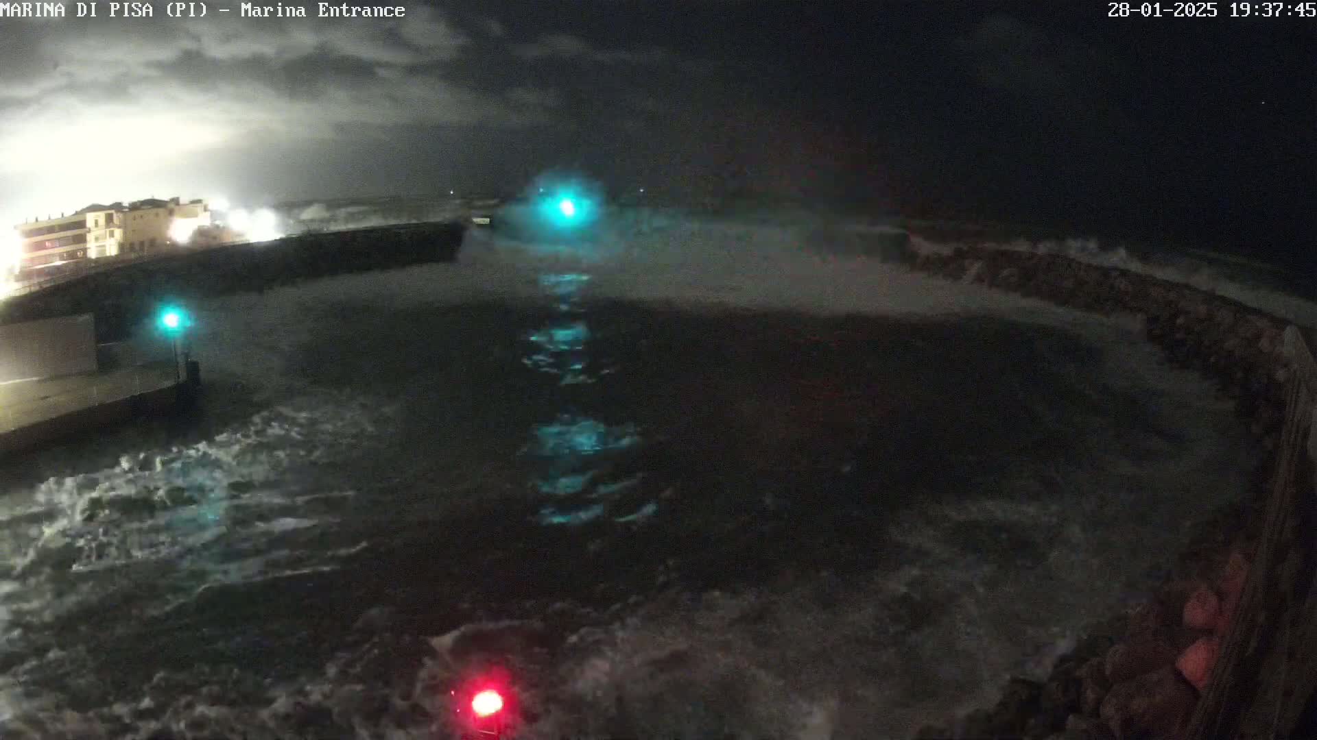 Large waves crash against a seawall at night, illuminated by several lights on the wall and in the water.