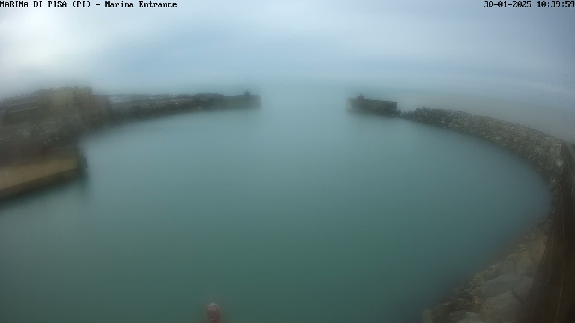 A foggy view of a harbor entrance with calm, murky green water enclosed by stone walls.