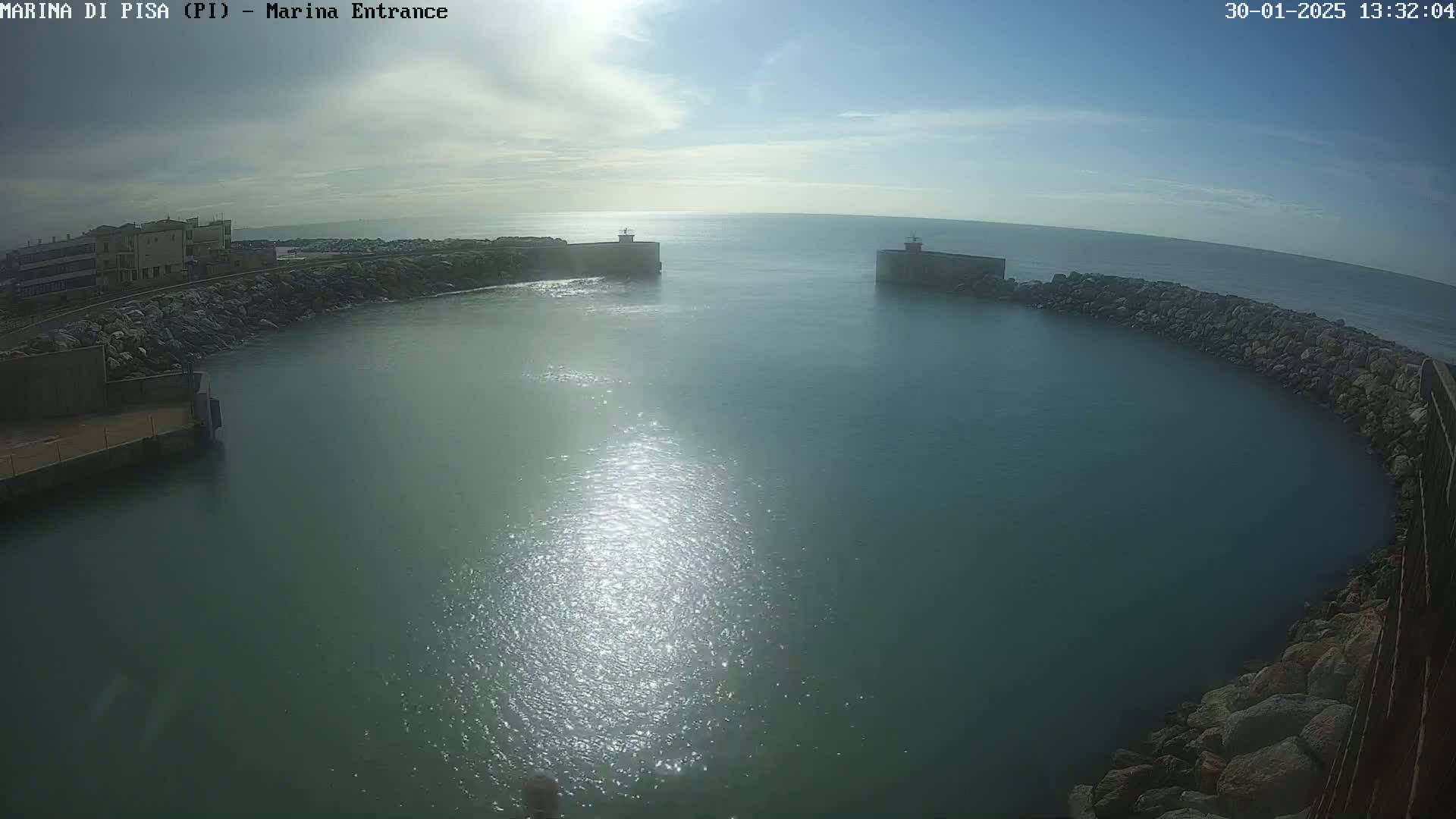 A mostly sunny day shows a calm harbor entrance, partially enclosed by rocky breakwaters and buildings.