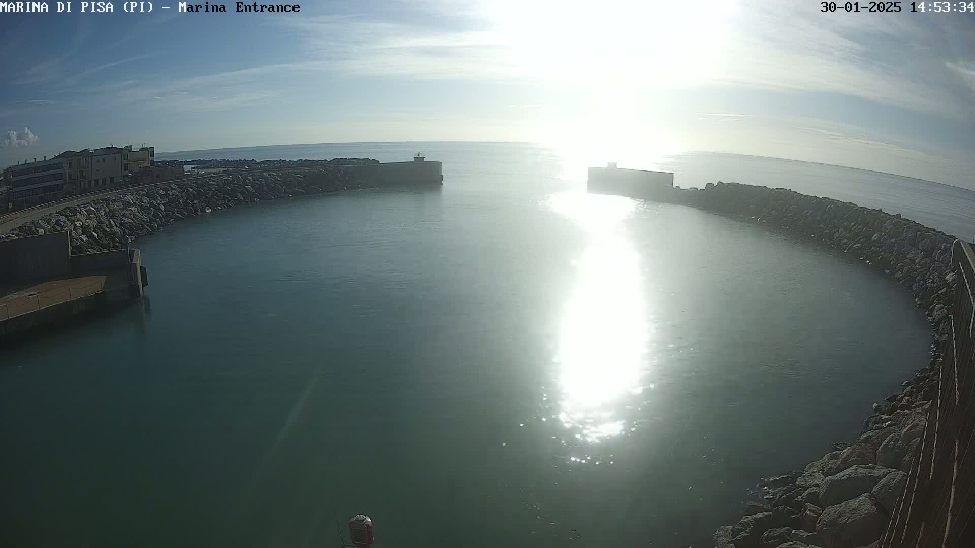A harbor entrance with calm, sunlit water is surrounded by rock walls, with buildings visible in the background under a mostly clear sky.