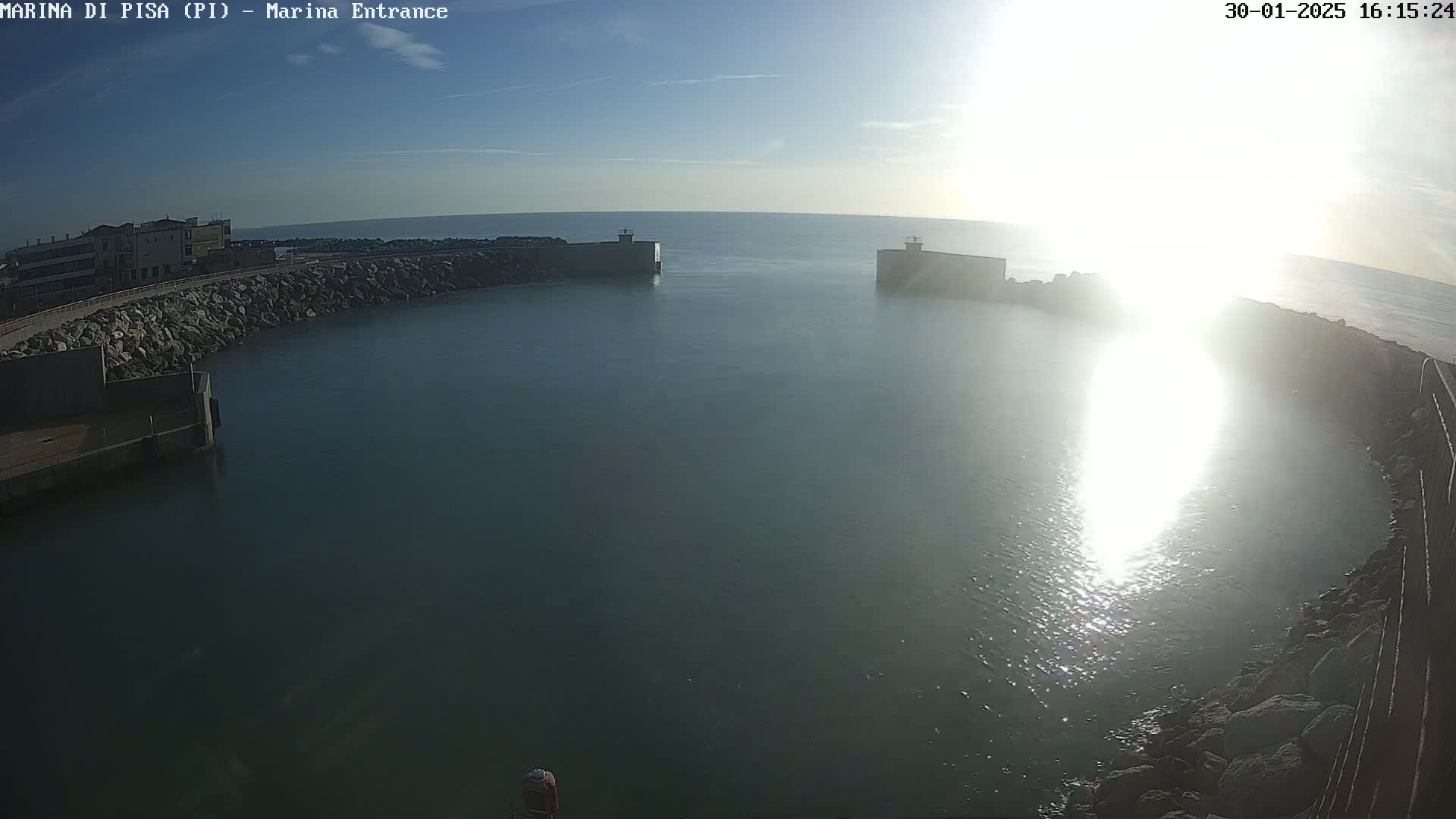 A harbor entrance with calm water is seen on a sunny day, with two breakwaters on either side of the entryway.