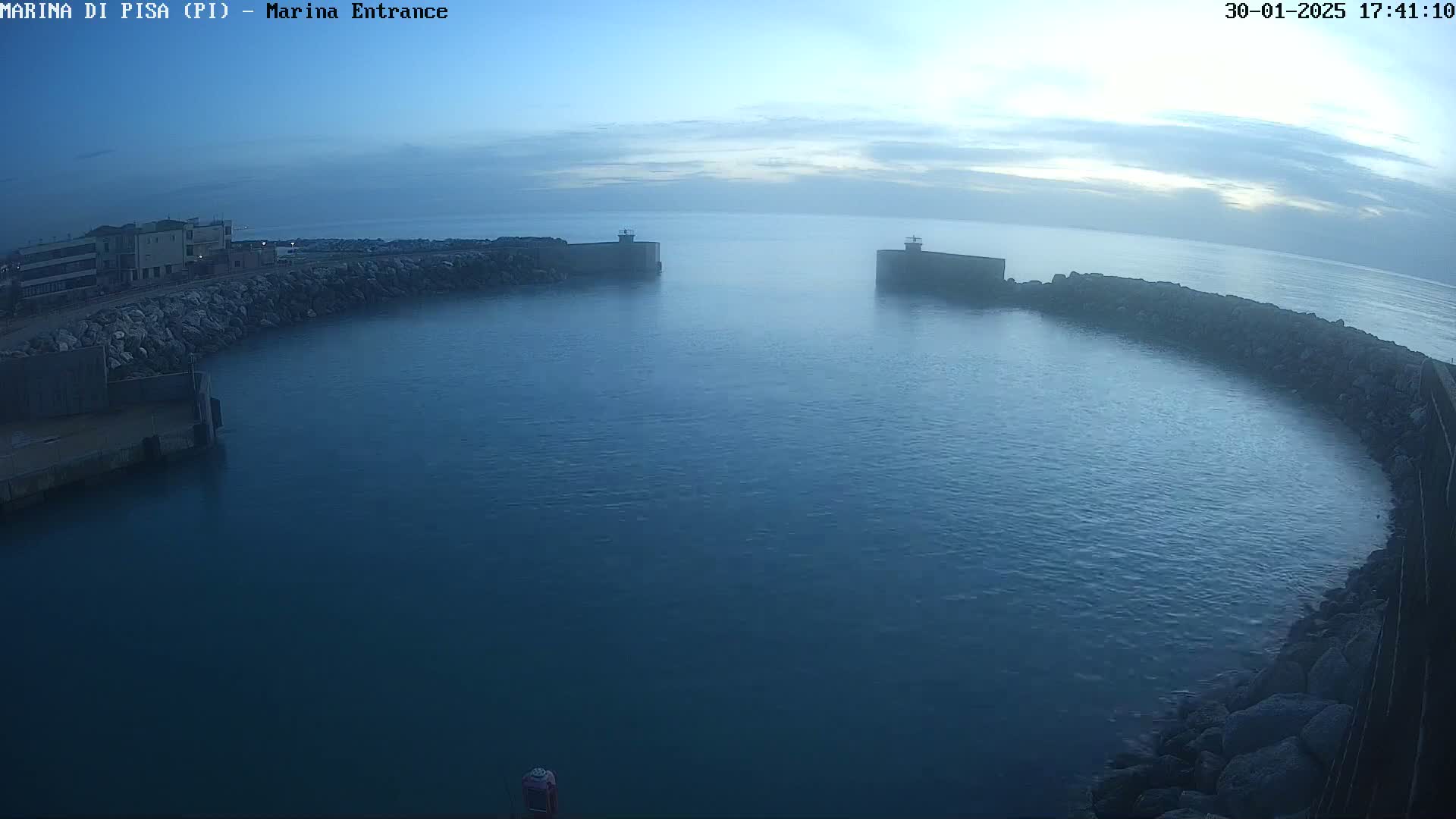 A calm, dark-blue body of water is enclosed by a curved stone wall, with a building visible on the left and a hazy sky above.