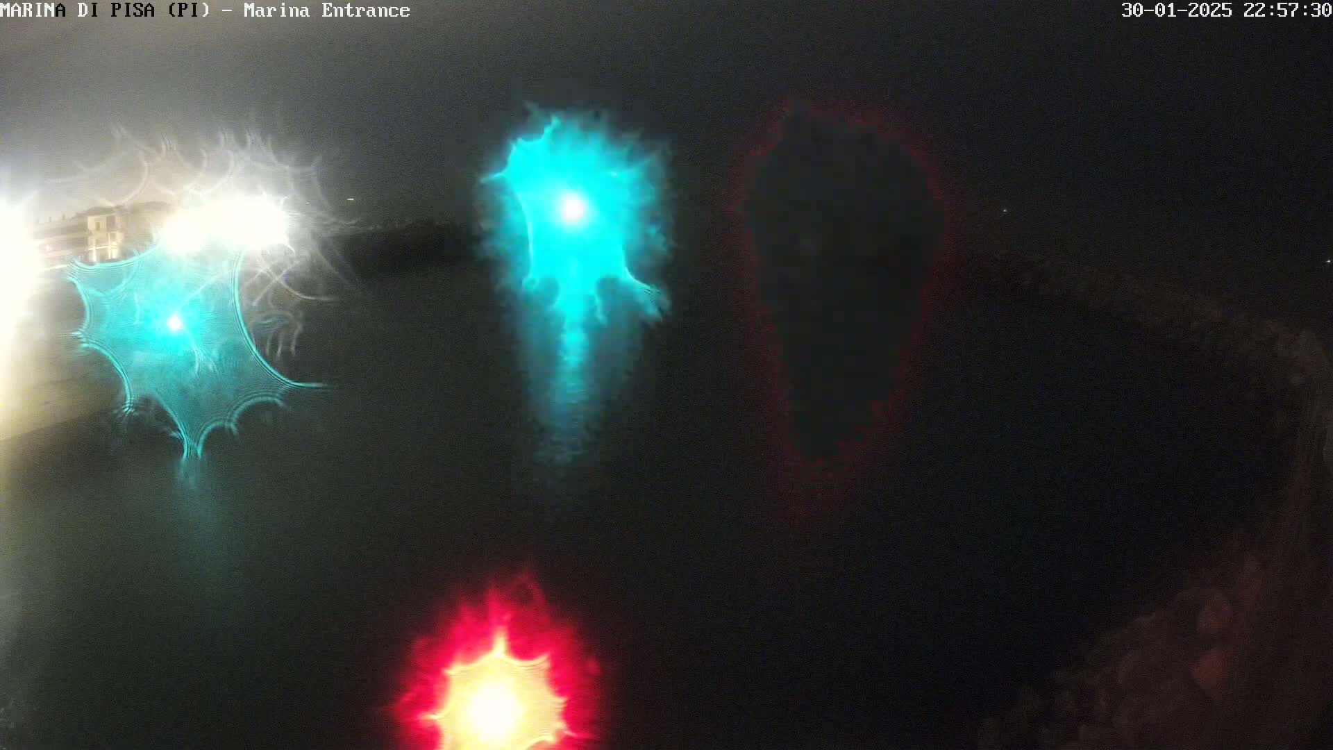 A nighttime view of a dark body of water with distorted reflections of lights in teal, white, and red, appearing as blurry streaks and glows.