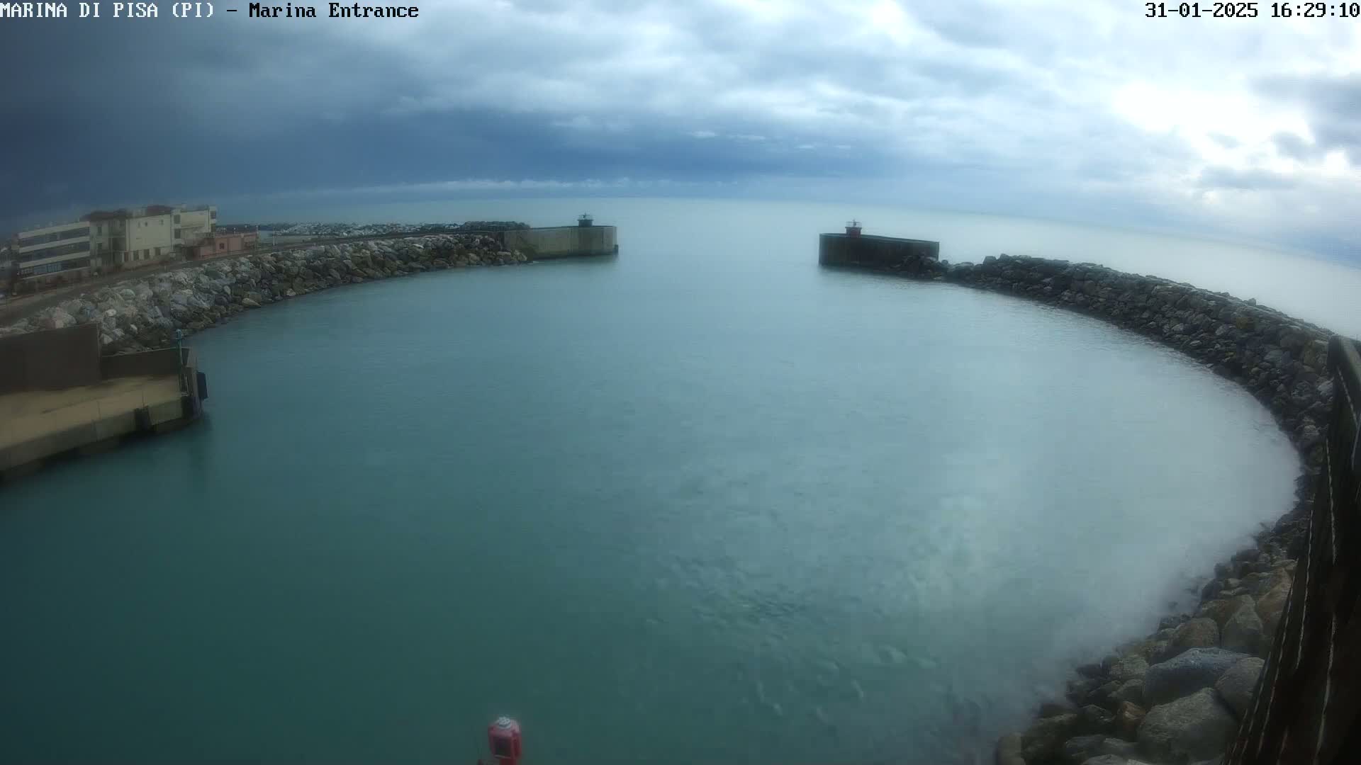 A calm, teal-colored harbor is seen under an overcast sky, enclosed by rock walls and buildings on one side.