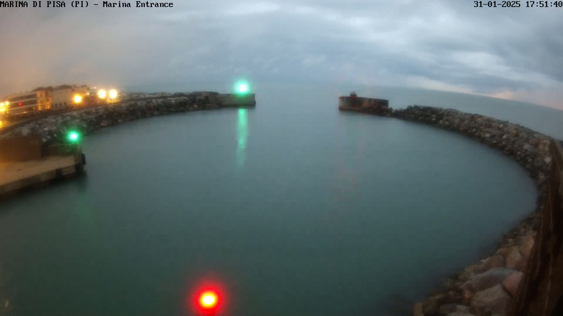 A calm, overcast harbor entrance is enclosed by stone walls with lights illuminating the water at dusk.
