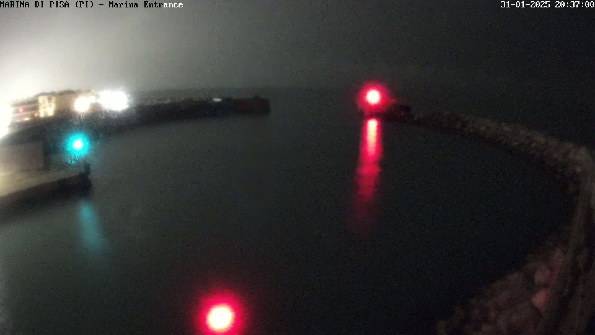 A nighttime view of a harbor entrance with two red navigation lights and one green navigation light visible on the water, under hazy or foggy conditions.