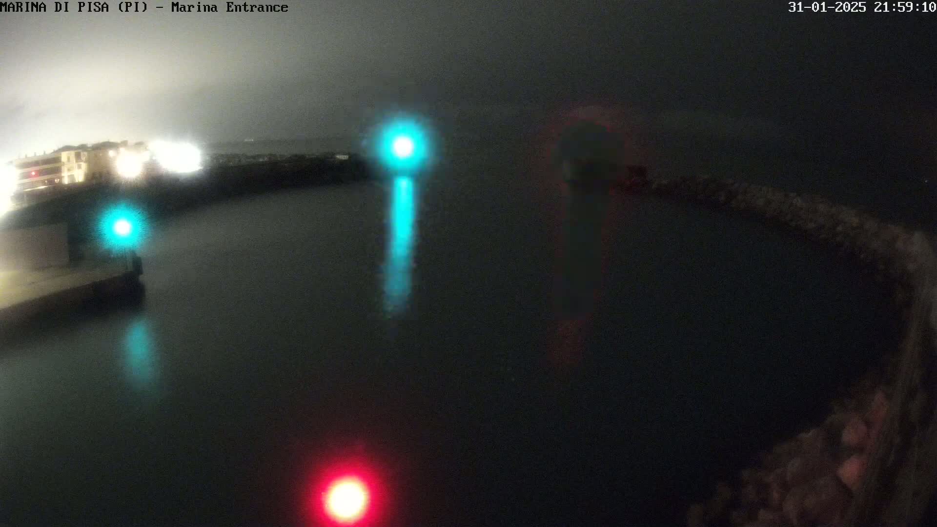 A nighttime view of a dark body of water with lights reflecting on the surface, illuminated by a teal and a red light on either side of a dark structure, under a foggy sky.