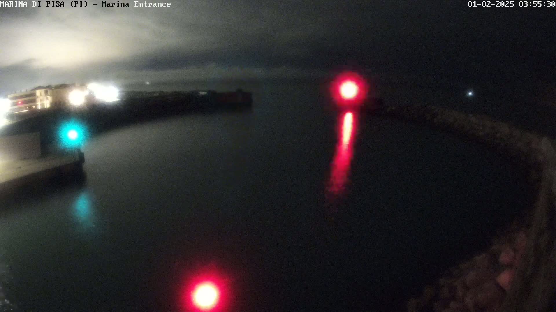 A nighttime view of a dark harbor with red and green navigation lights reflecting on the water under an overcast sky.